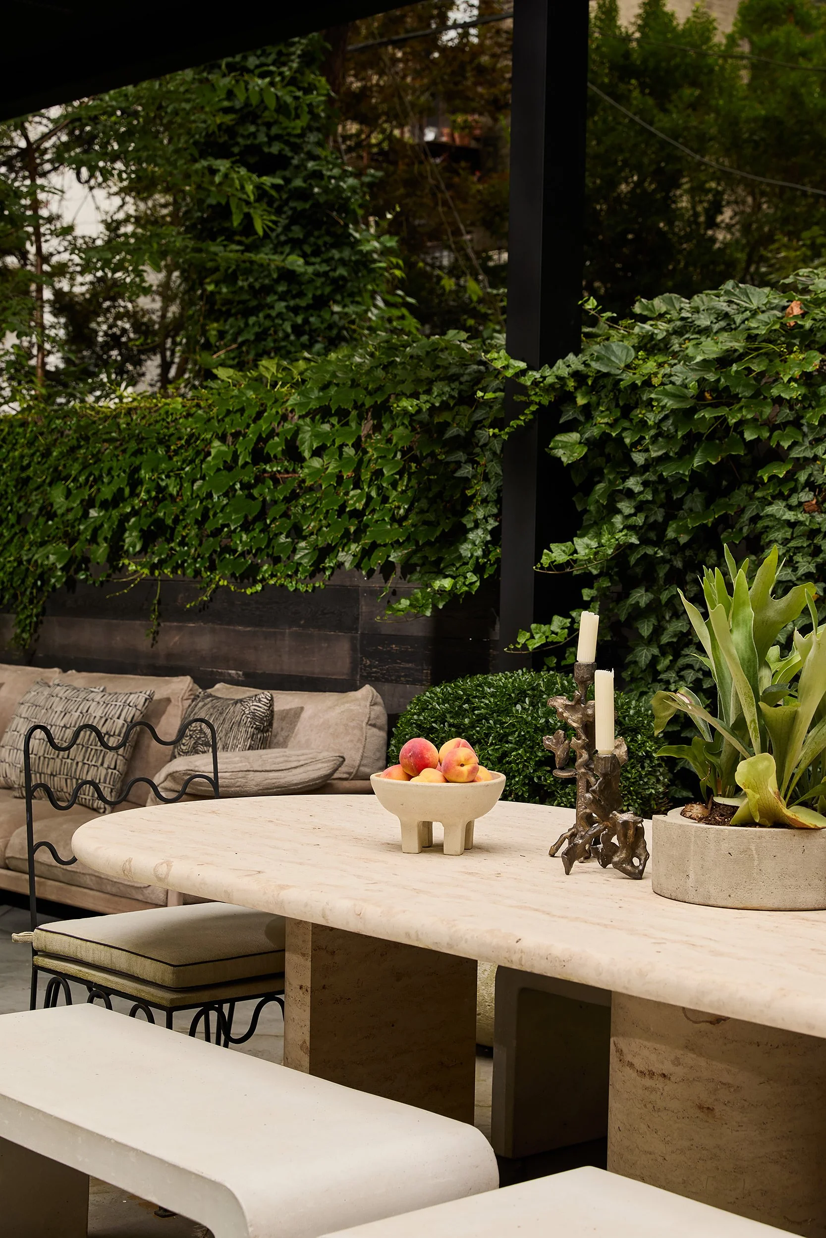 Outdoor patio with beige stone dining table, black metal chairs, a beige sofa with patterned cushions, and lush green foliage in the background.