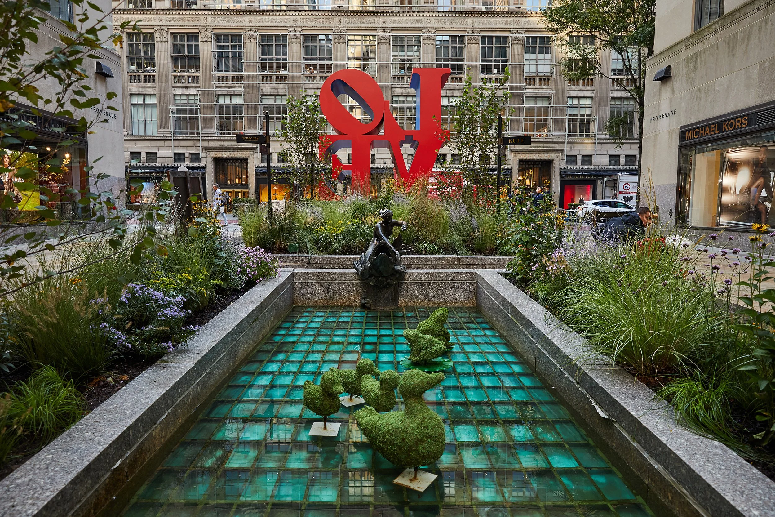 Urban park with fountain featuring animal topiary, large red LOVE sculpture, and surrounding plants, city buildings, and retail shops.