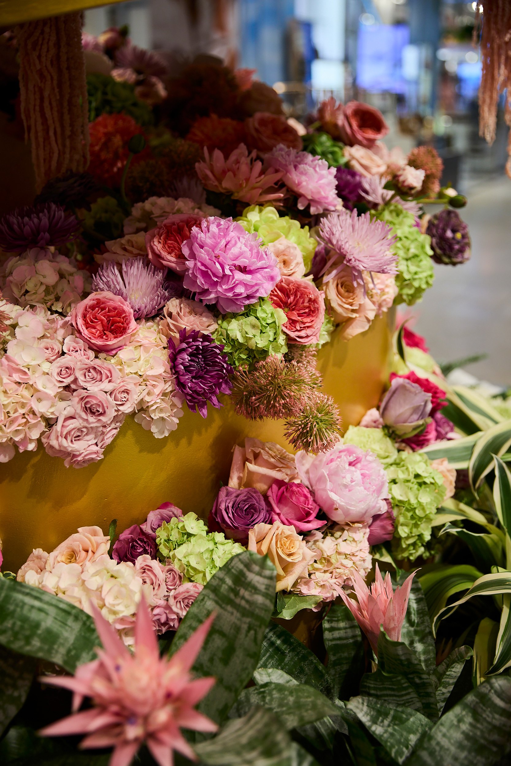 A colorful display of various flowers, including roses, hydrangeas, and lilies, arranged in a yellow container in a flower shop.