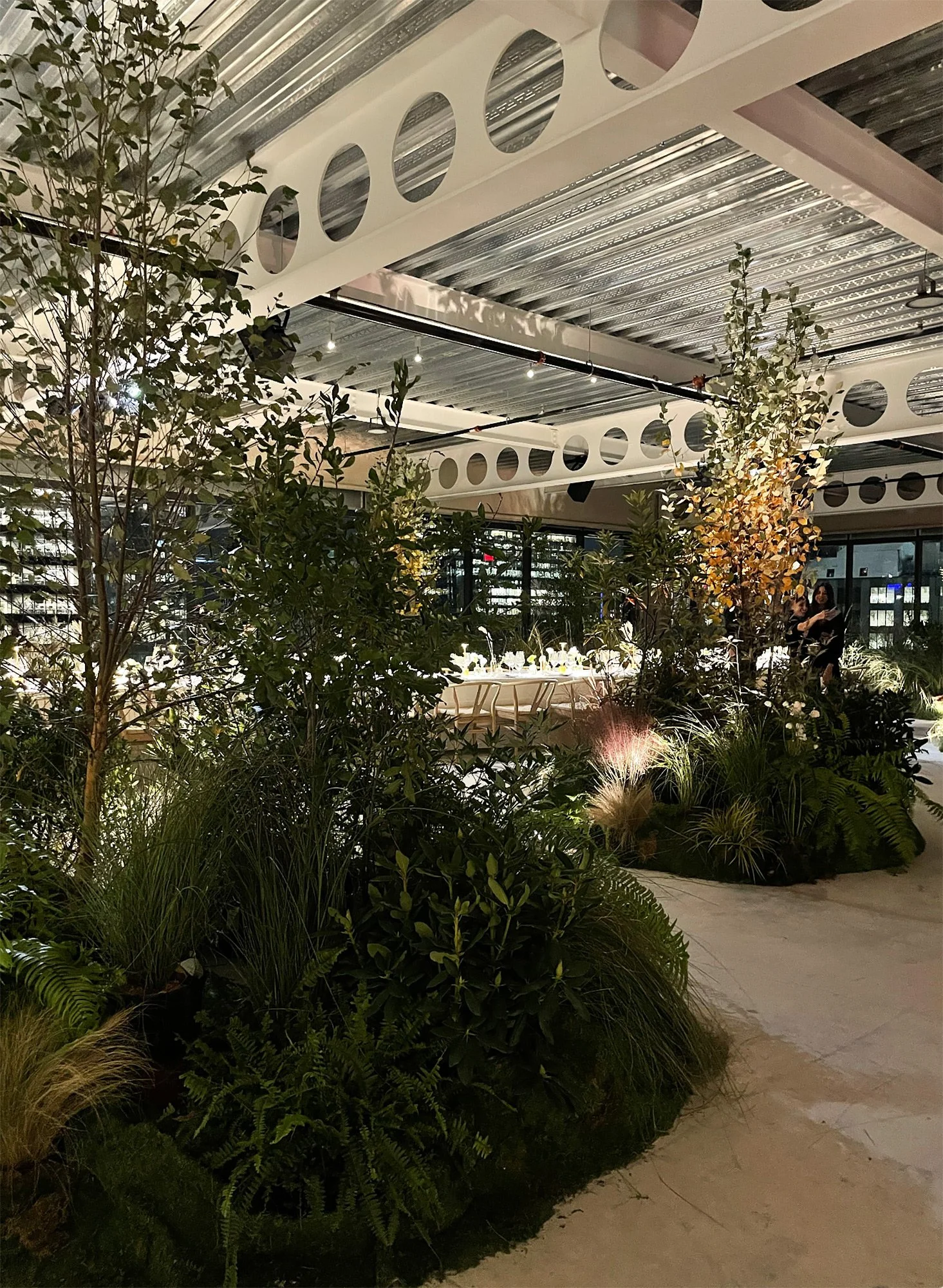 Indoor event space decorated with lush green plants and trees, with a long table set for a formal dinner in the background, featuring elegant tableware and chairs.