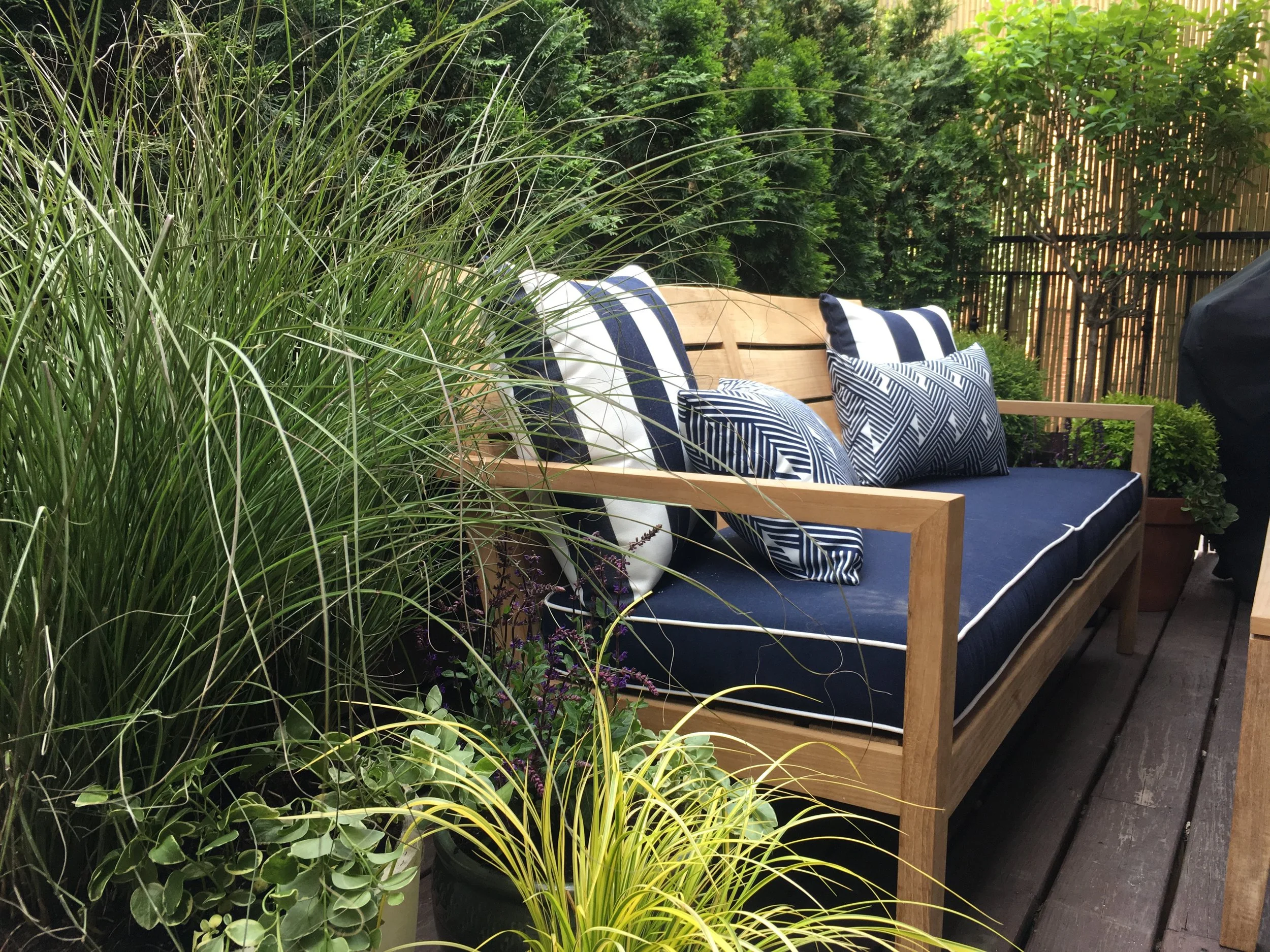 A wooden outdoor bench with dark blue cushions and black and white striped pillows, surrounded by lush green plants and trees in a backyard or garden setting.