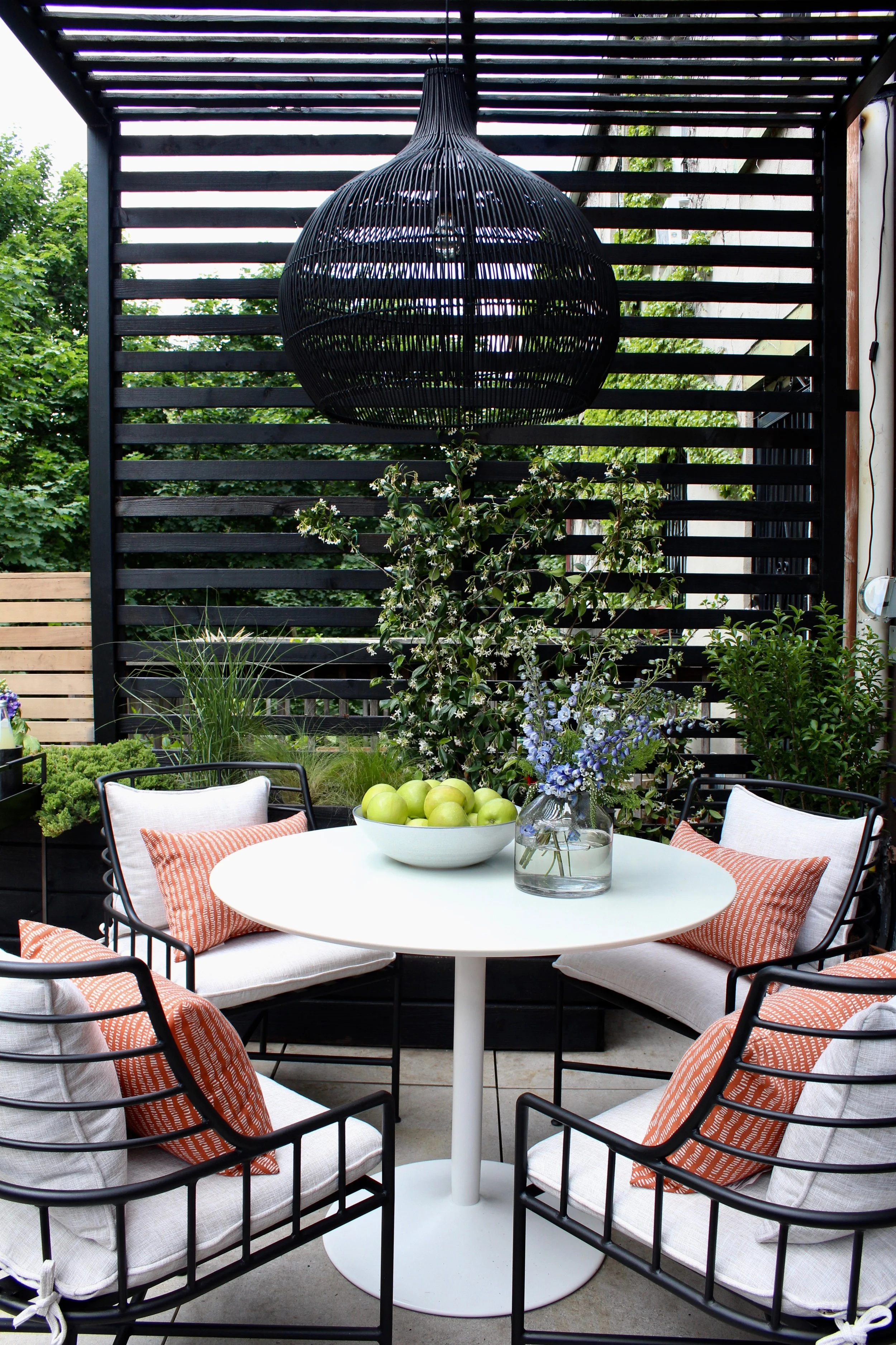 Outdoor patio with a round white table surrounded by black metal chairs with white cushions and orange striped pillows. On the table, a white bowl of green apples and a glass vase with blue and white flowers. A black wooden slat privacy screen with greenery and trees in the background. A large black woven pendant light hanging above.