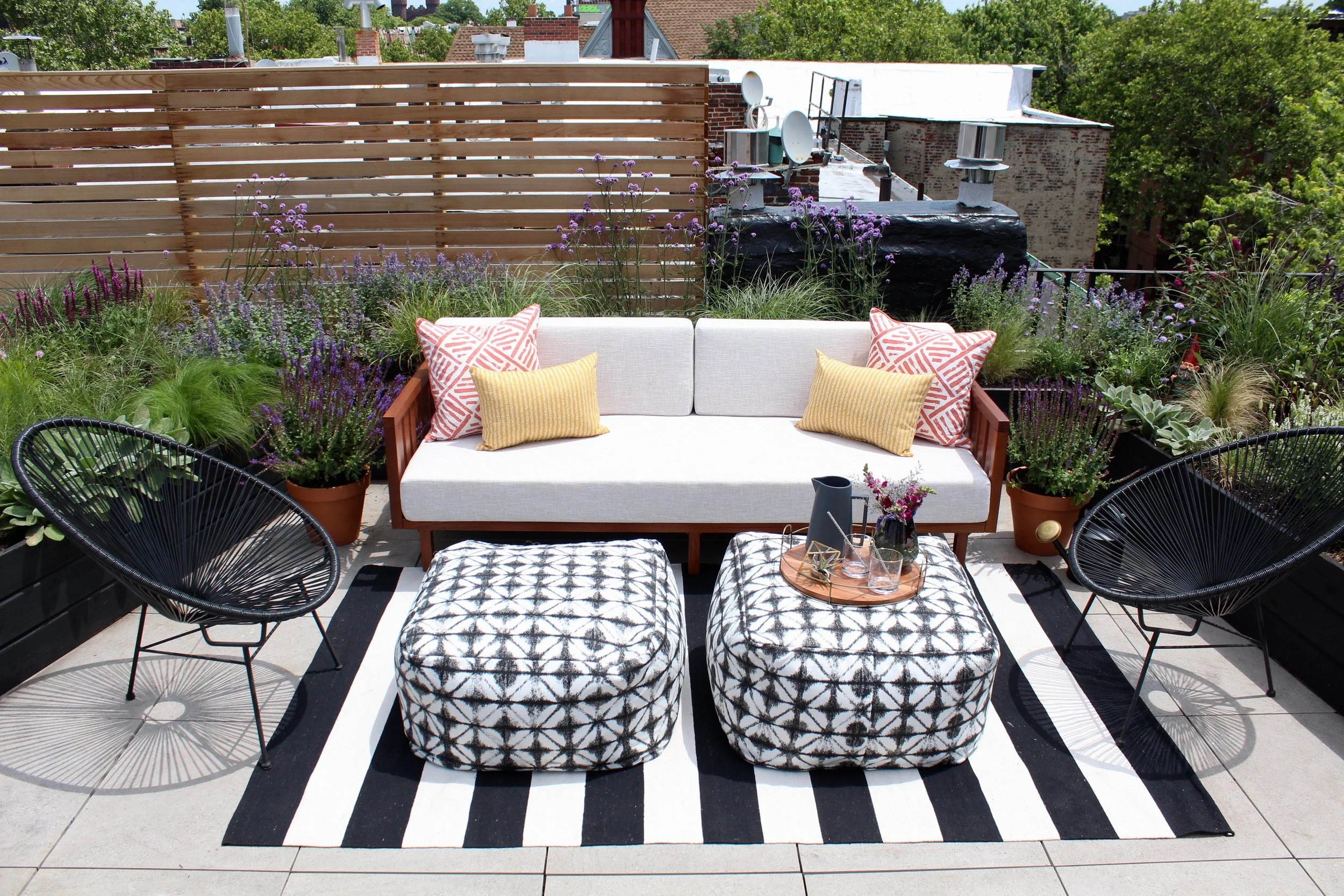 Outdoor rooftop patio with white cushioned sofa, black lounge chairs, patterned ottomans, and potted plants. The area is decorated with a black and white striped rug, and surrounded by a wooden privacy fence and lush trees, with buildings visible in the background.
