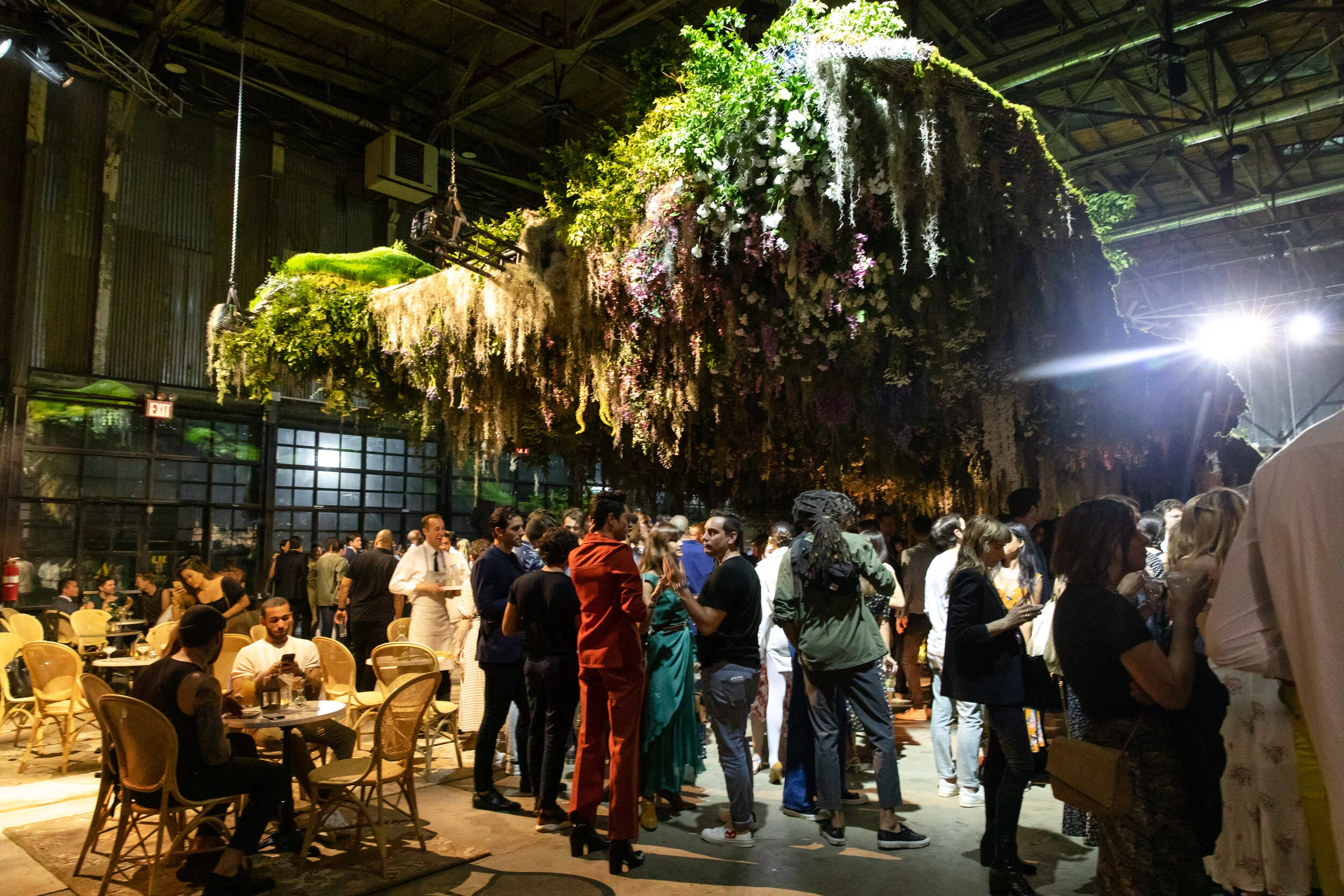 An indoor event with many people walking and sitting at tables. The ceiling is decorated with hanging greenery and flowers, creating a lush environment.