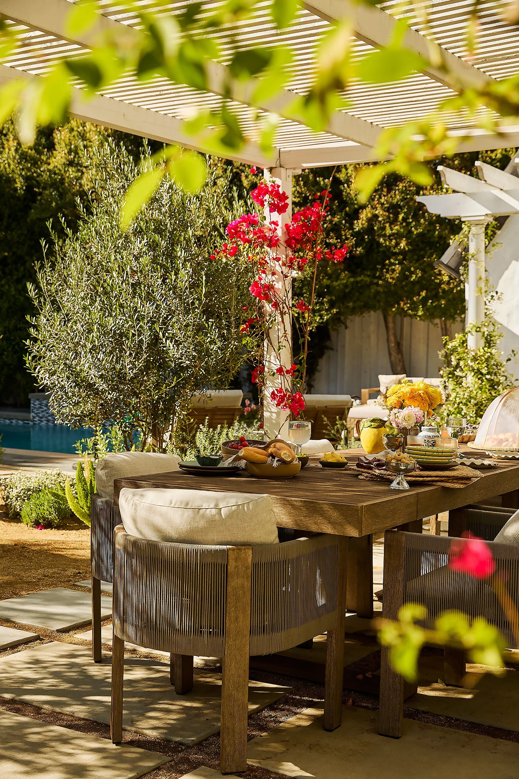 A backyard dining area with a wooden table set with food, drinks, and flowers, surrounded by modern chairs. There's a white pergola overhead, lush greenery, and a swimming pool in the background.