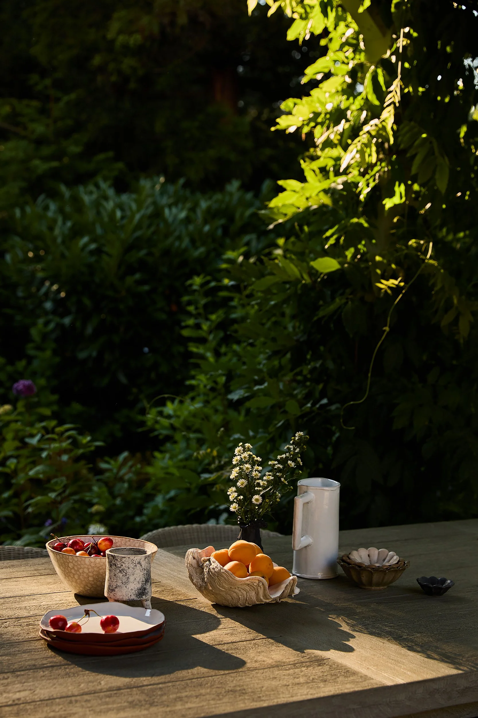 Outdoor wooden table with bowls of cherries and apricots, a white mug, a small vase of white flowers, and various small dishes, set in a lush garden with sunlight filtering through leaves.