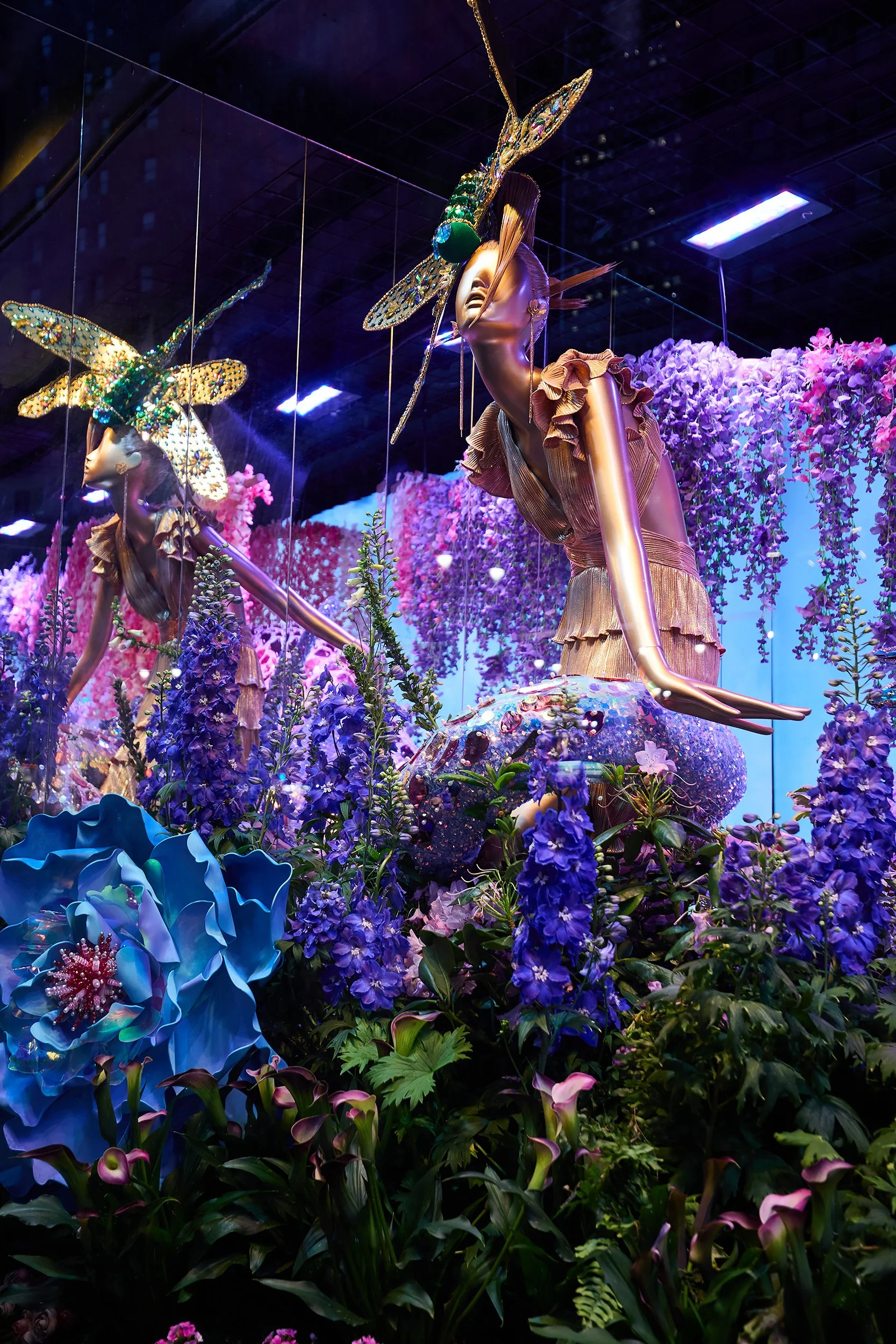 Display of mannequins with gold bodies and decorated with large insect headpieces made of beads and glitter, surrounded by purple and pink flowers in a vibrant, artistic setting.