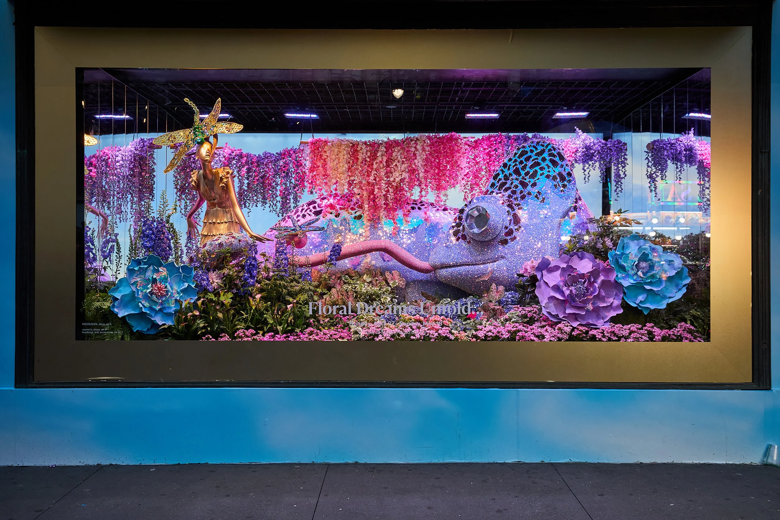 Display window with a floral installation featuring a pink and purple flower chameleon and a woman with a golden butterfly headpiece. The scene is decorated with large pink, purple, and blue flowers, and the backdrop is filled with hanging pink and purple flowers. The text 'Floral Dreams Unfold' is visible at the bottom.