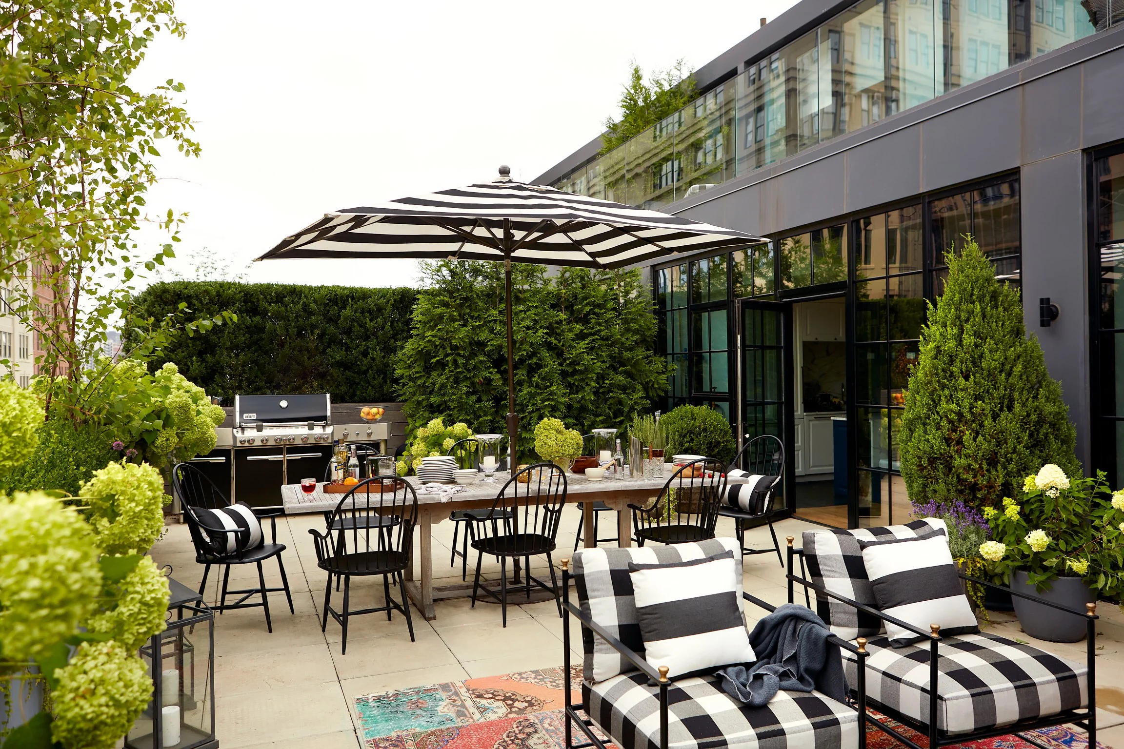 Outdoor patio with black and white decor, including striped cushions and pillows, a dining table with chairs, and a grill. Greenery surrounds the space, and there is a large black and white striped umbrella overhead.