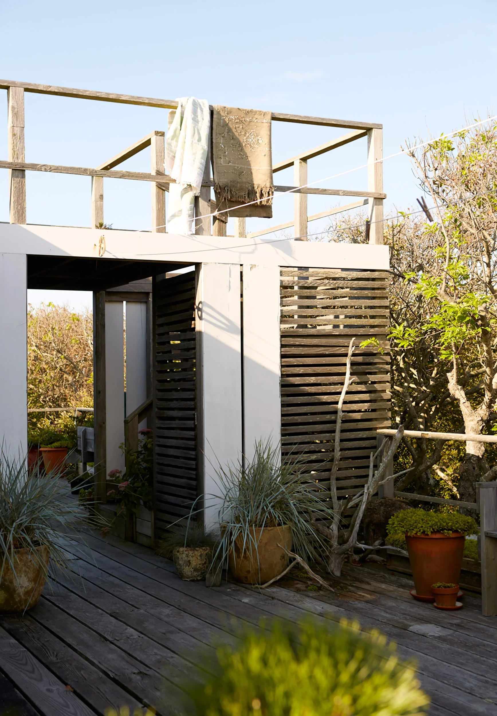 Outdoor rooftop patio with potted plants, a wooden deck, and a structure with wooden panels and white walls. Laundry towels hang on a railing above.