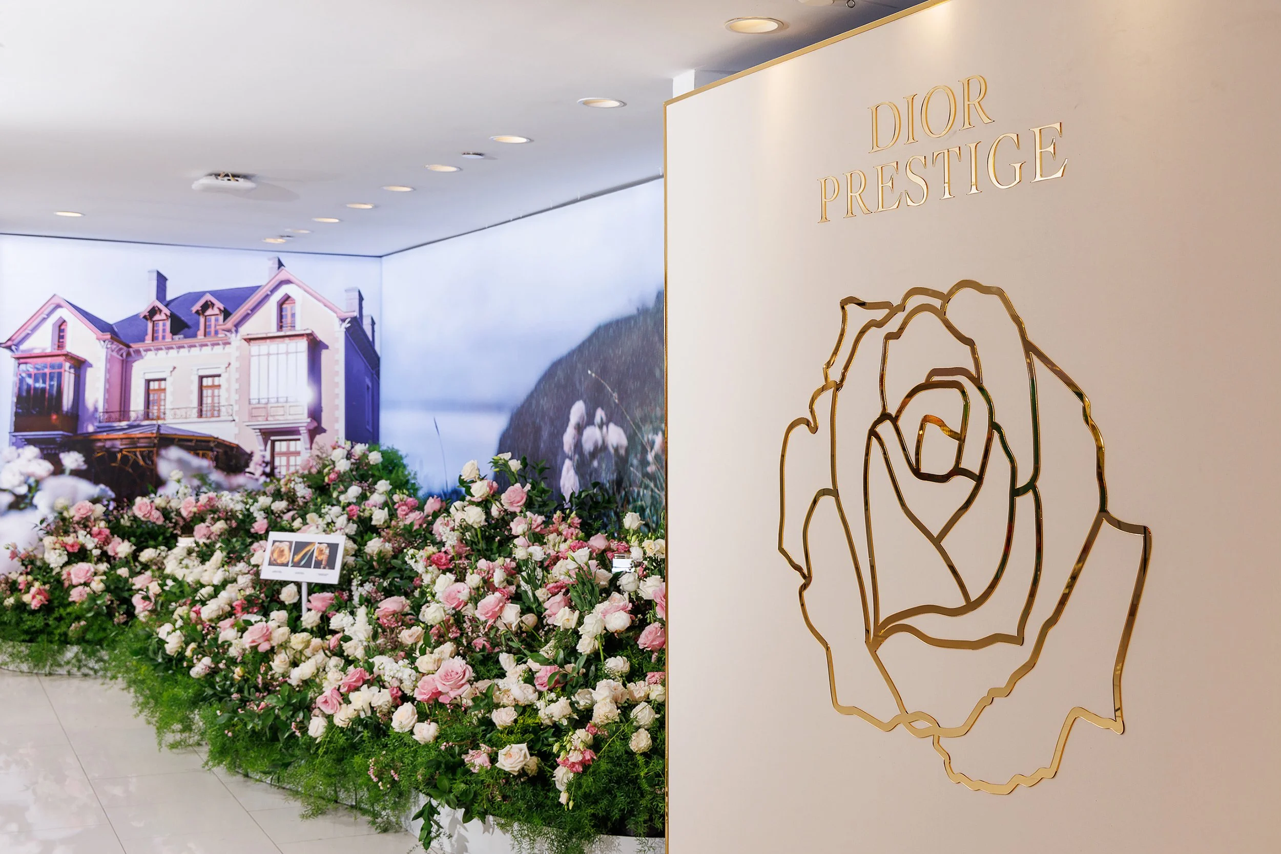 Display at Dior Prestige showcasing pink and white flowers with a backdrop of a house painting and a white wall with a gold outline rose and Dior Prestige logo.