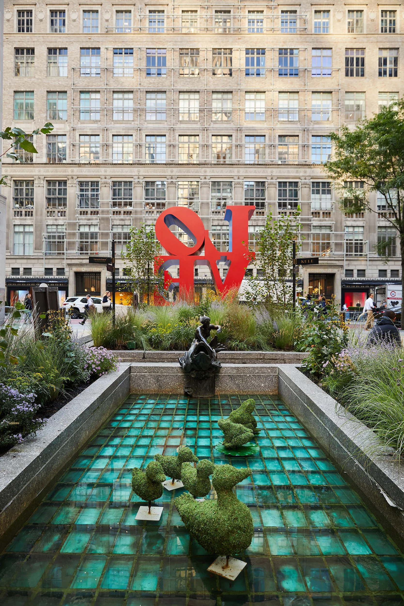 Public park with a large red sculpture of the word "LOVE" on a city street, surrounded by greenery, flowers, and small topiary animals in a water feature, with buildings and pedestrians in the background.