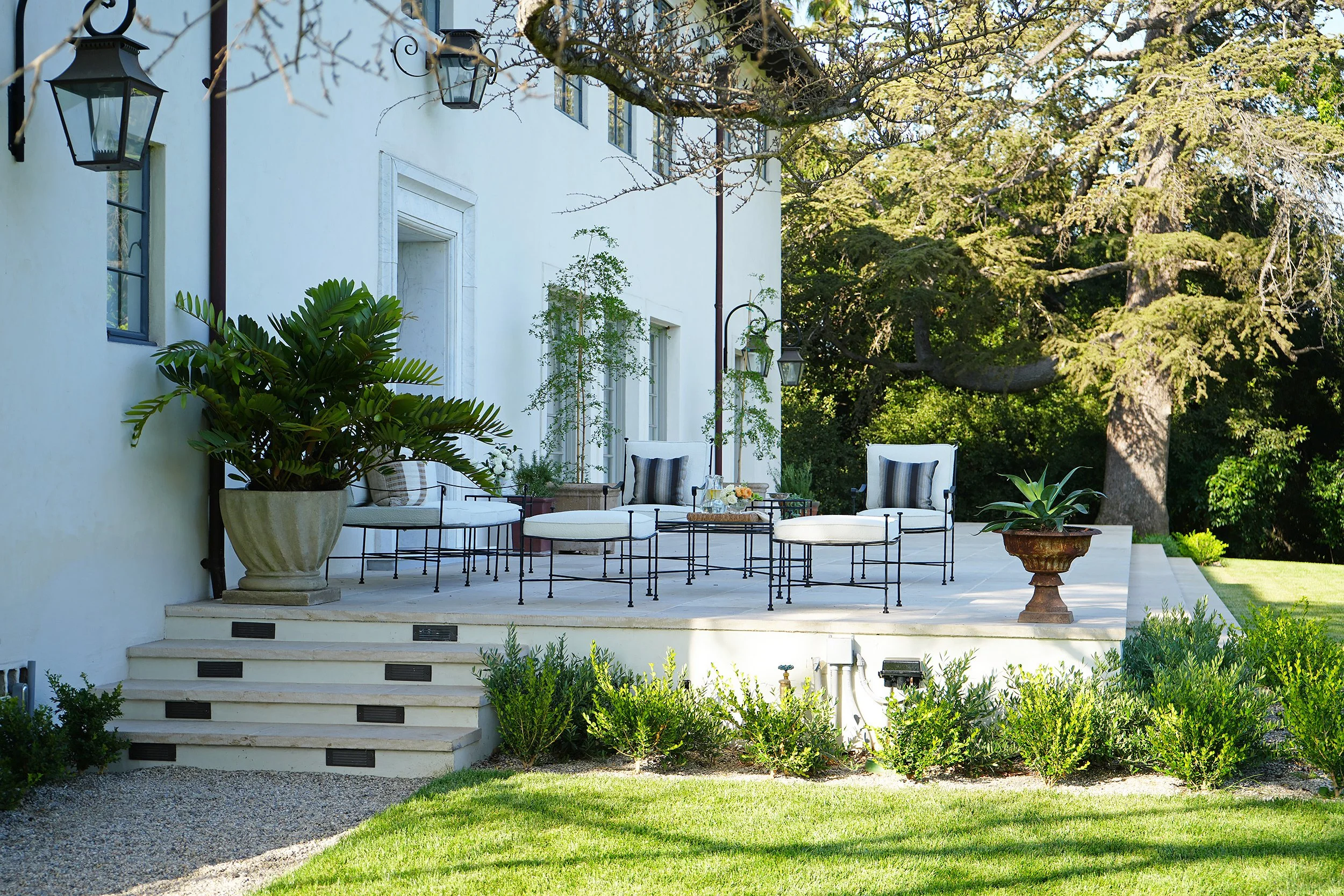 Outdoor patio area with white chairs, green plants in large pots, and a large tree in a lush garden.
