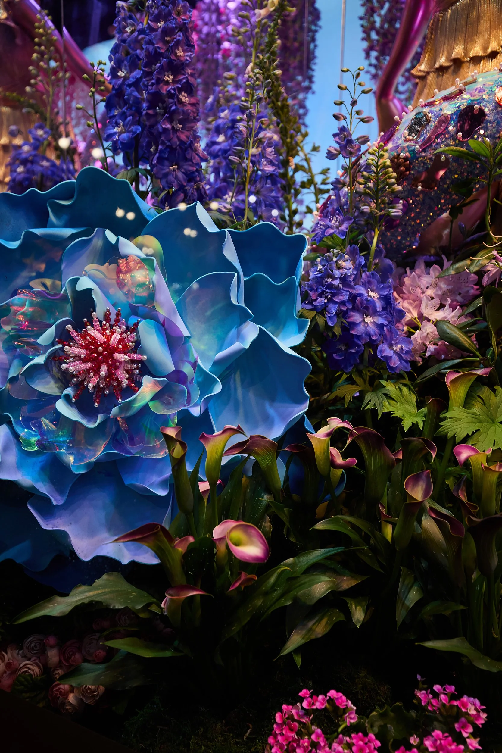 Colorful artificial flowers and plants in vibrant purple, pink, and blue hues, with large blue petal structures and purple-flowered tall stalks.