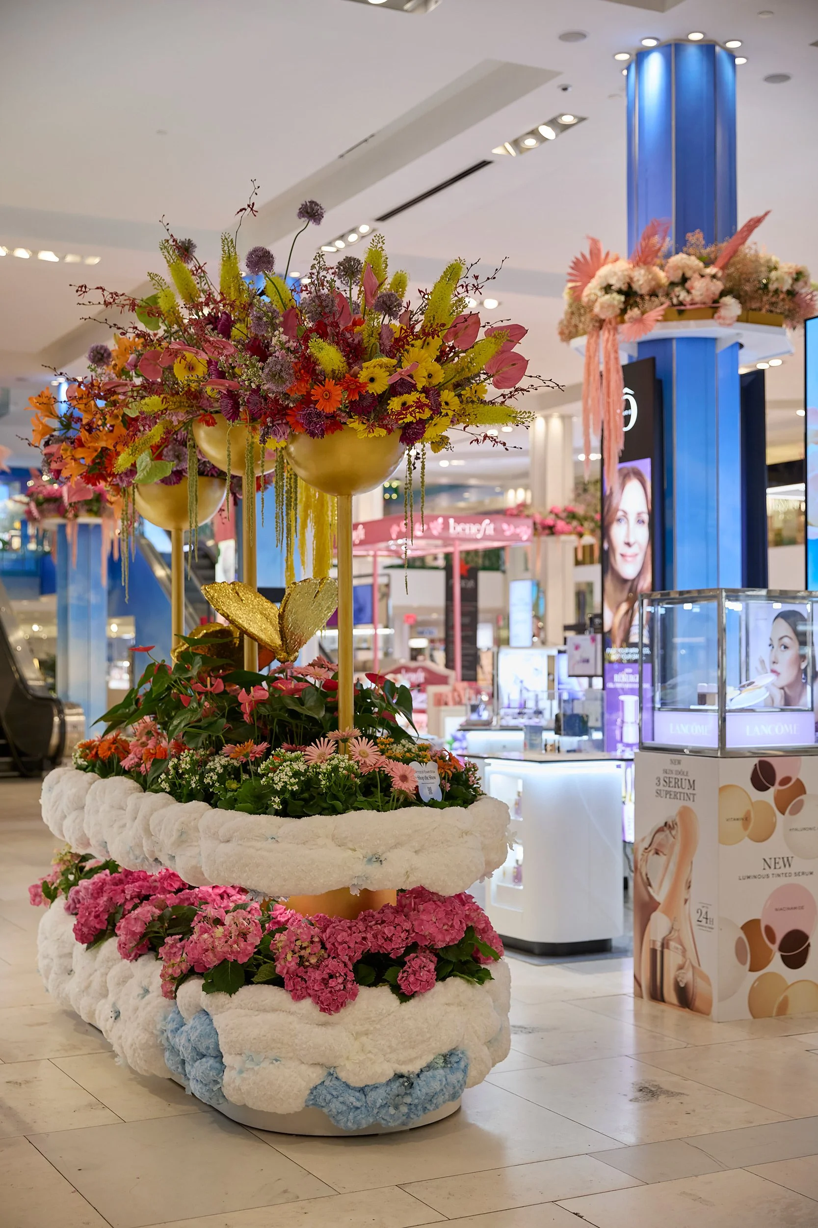 Colorful floral display with multiple layers of flowers and greenery inside a shopping mall.