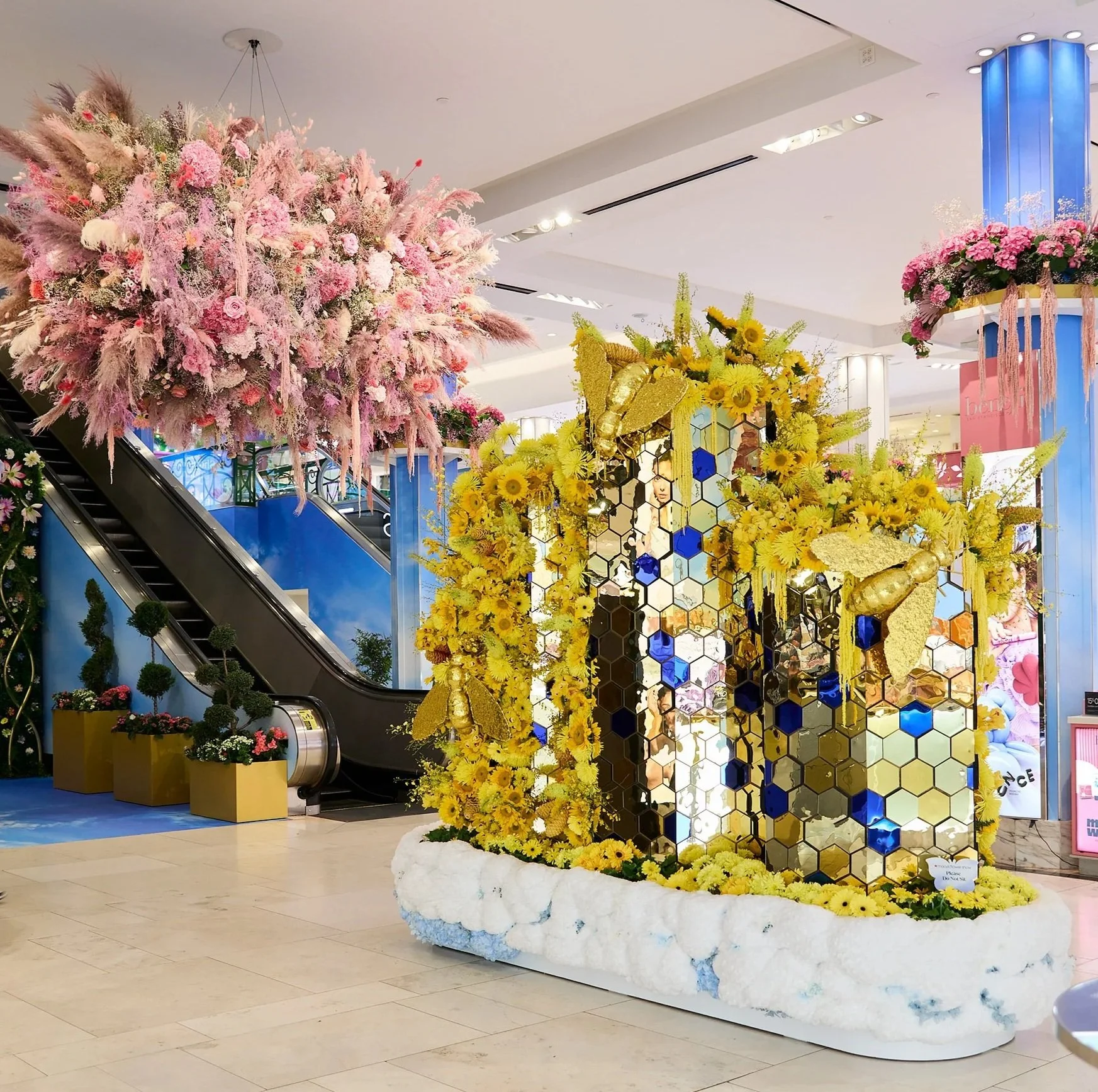 Decorative floral display featuring yellow flowers and gold butterflies with a honeycomb-patterned backdrop in a shopping mall.