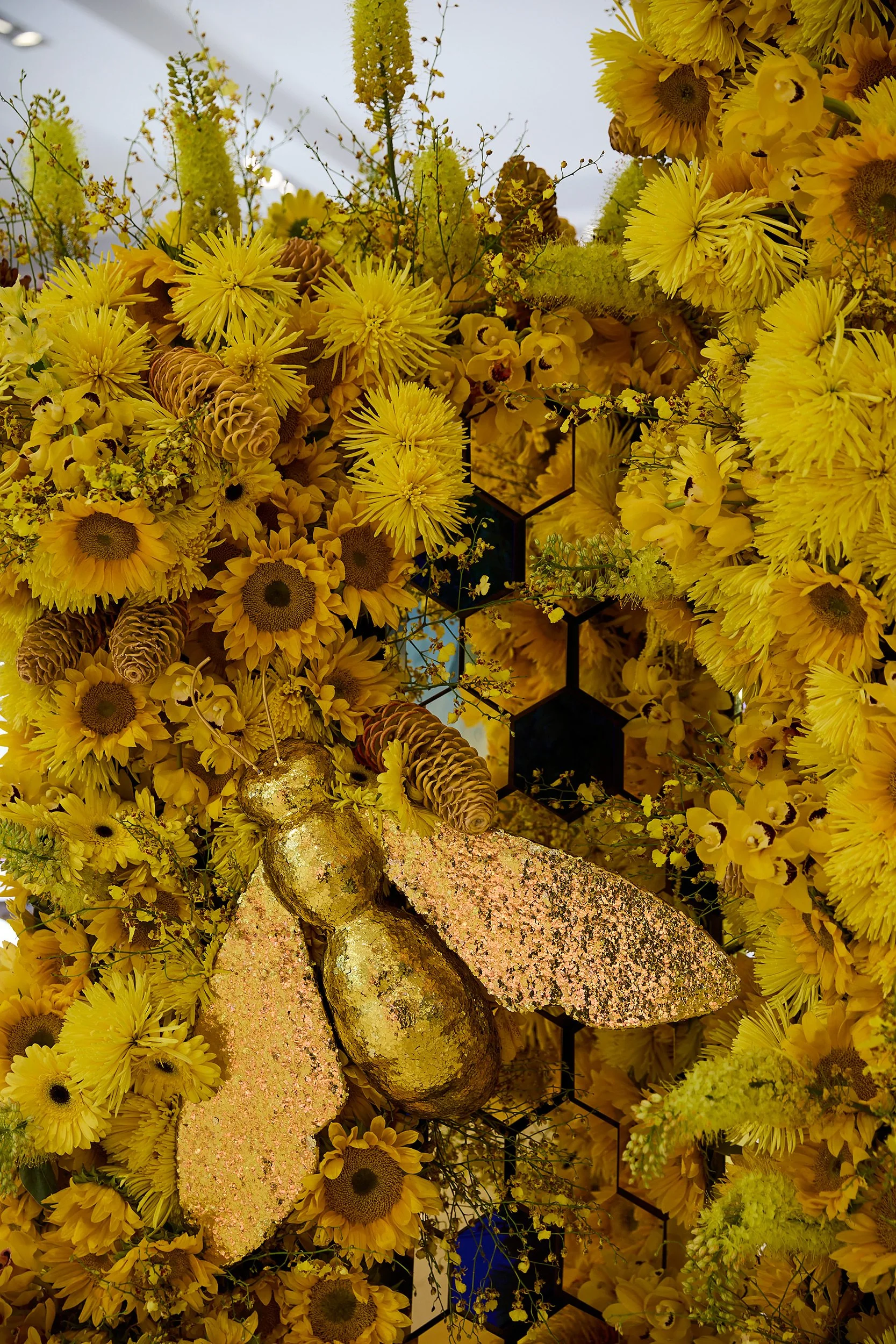 A large, elaborate floral arrangement featuring yellow flowers, pinecones, and a metallic bee ornament with gold wings.