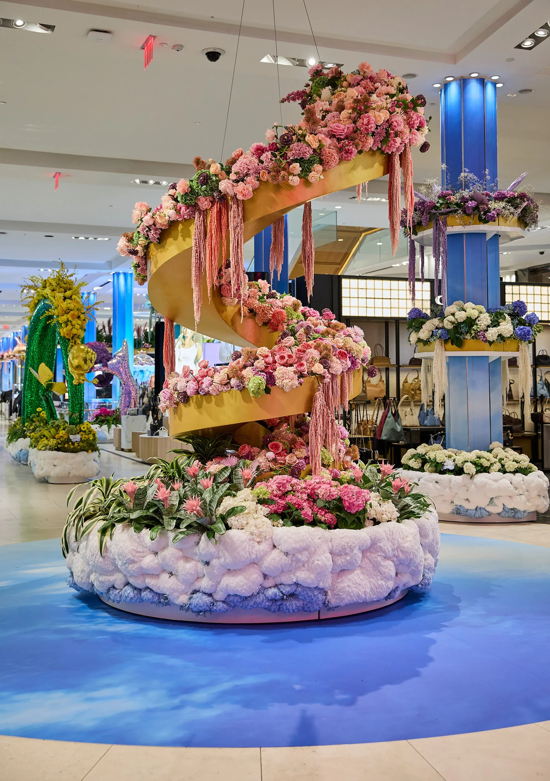 Indoor display with a spiral floral sculpture decorated with pink, white, and green flowers, surrounded by a tie-dye blue base that resembles water.