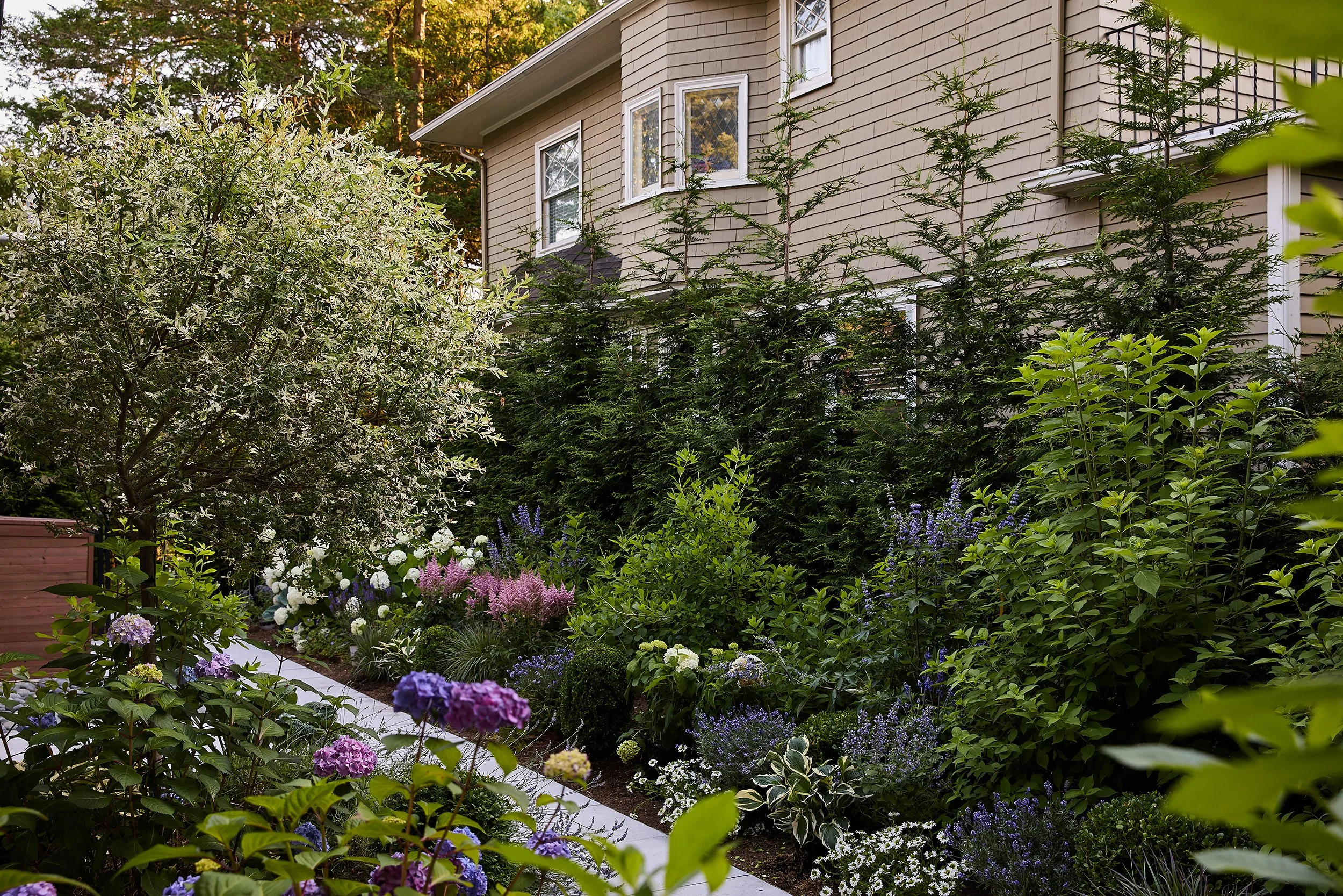 A lush backyard garden with flowering plants, shrubs, and small trees along the side of a house with beige siding and white trim.