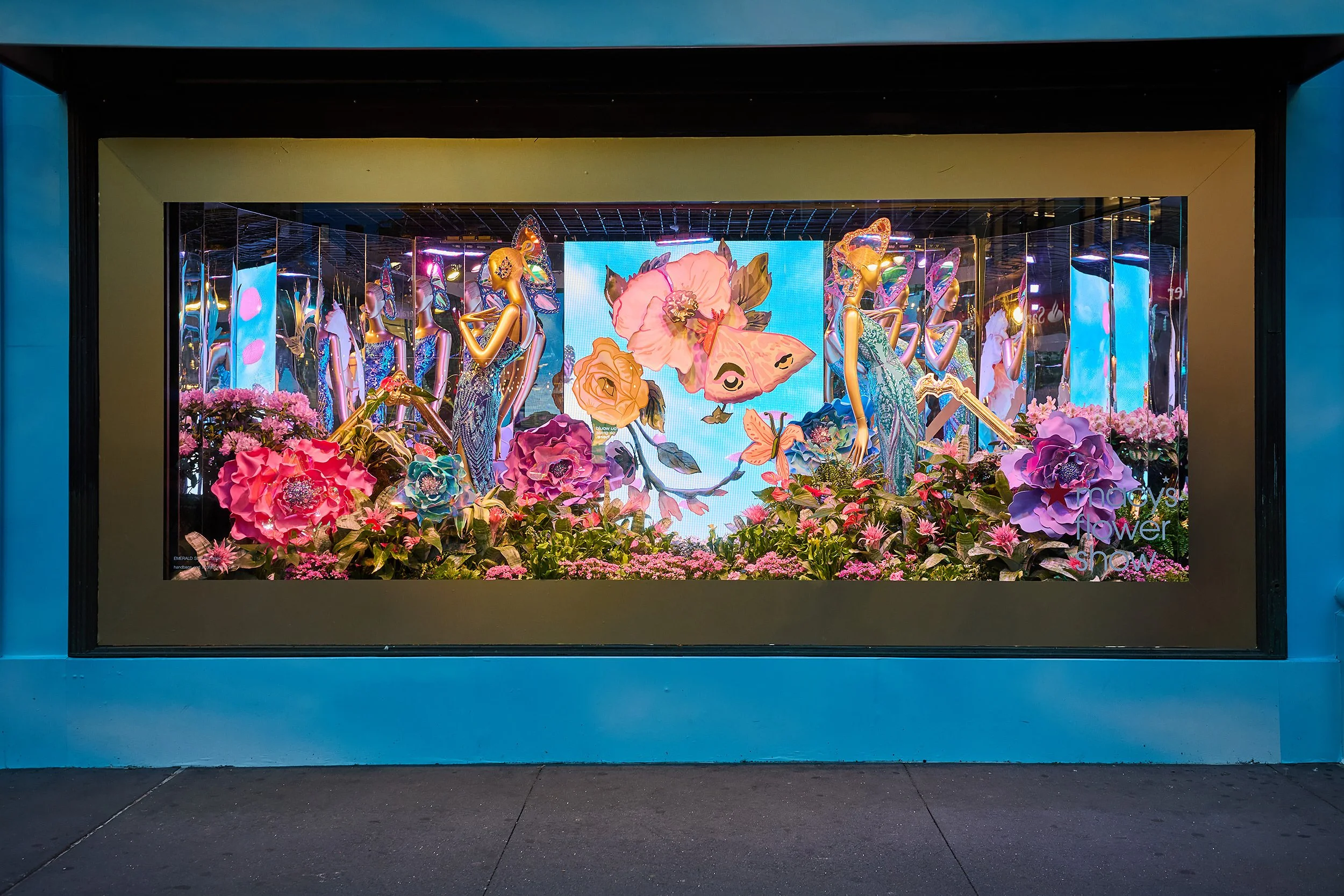 A storefront display window featuring a colorful floral and fashion-themed scene with mannequins dressed in vibrant outfits, surrounded by oversized flowers and butterflies, with a digital butterfly projection in the background.