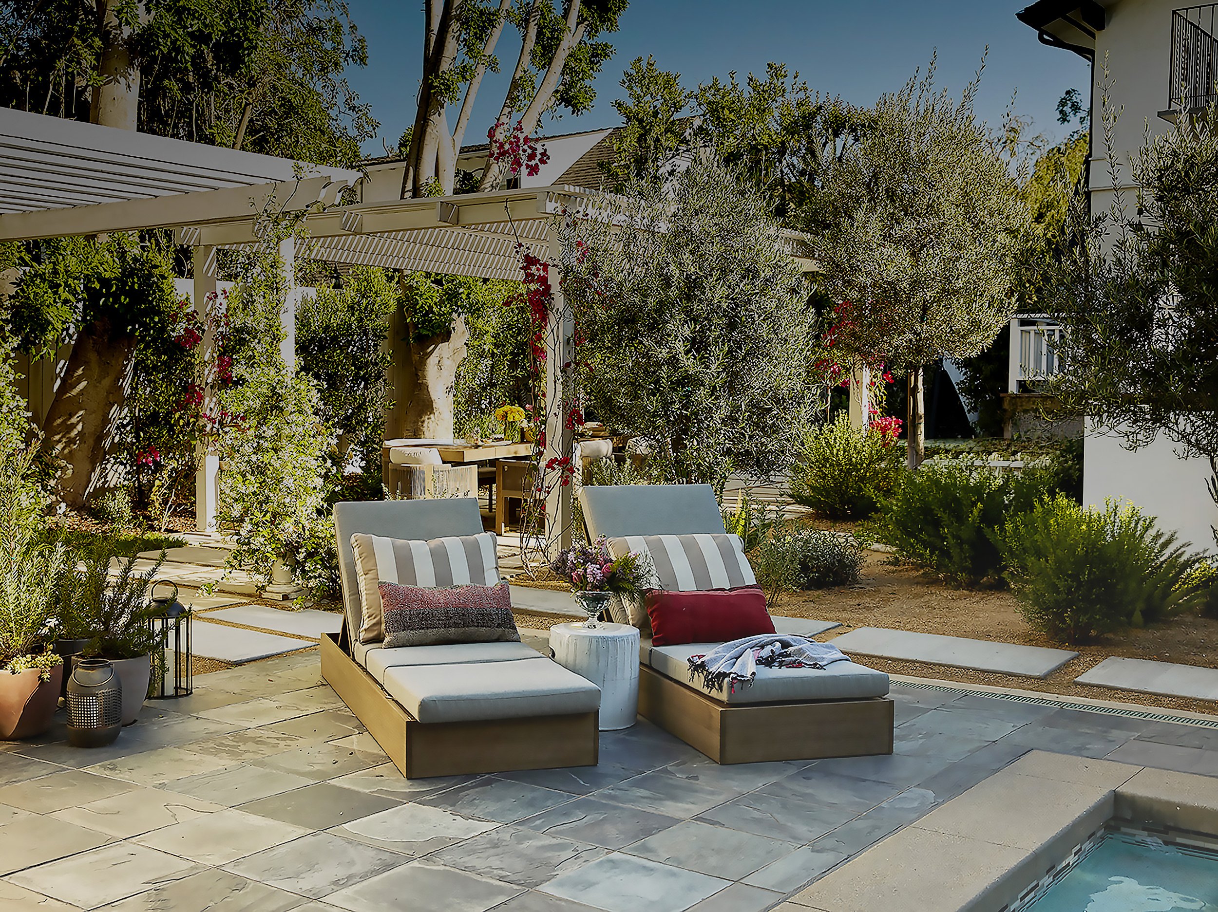 A cozy outdoor patio area with lounge chairs, a small round table with flowers, surrounded by lush greenery, trees, and bushes. There are cushions and a blanket on the chairs, with a house visible on the right side.