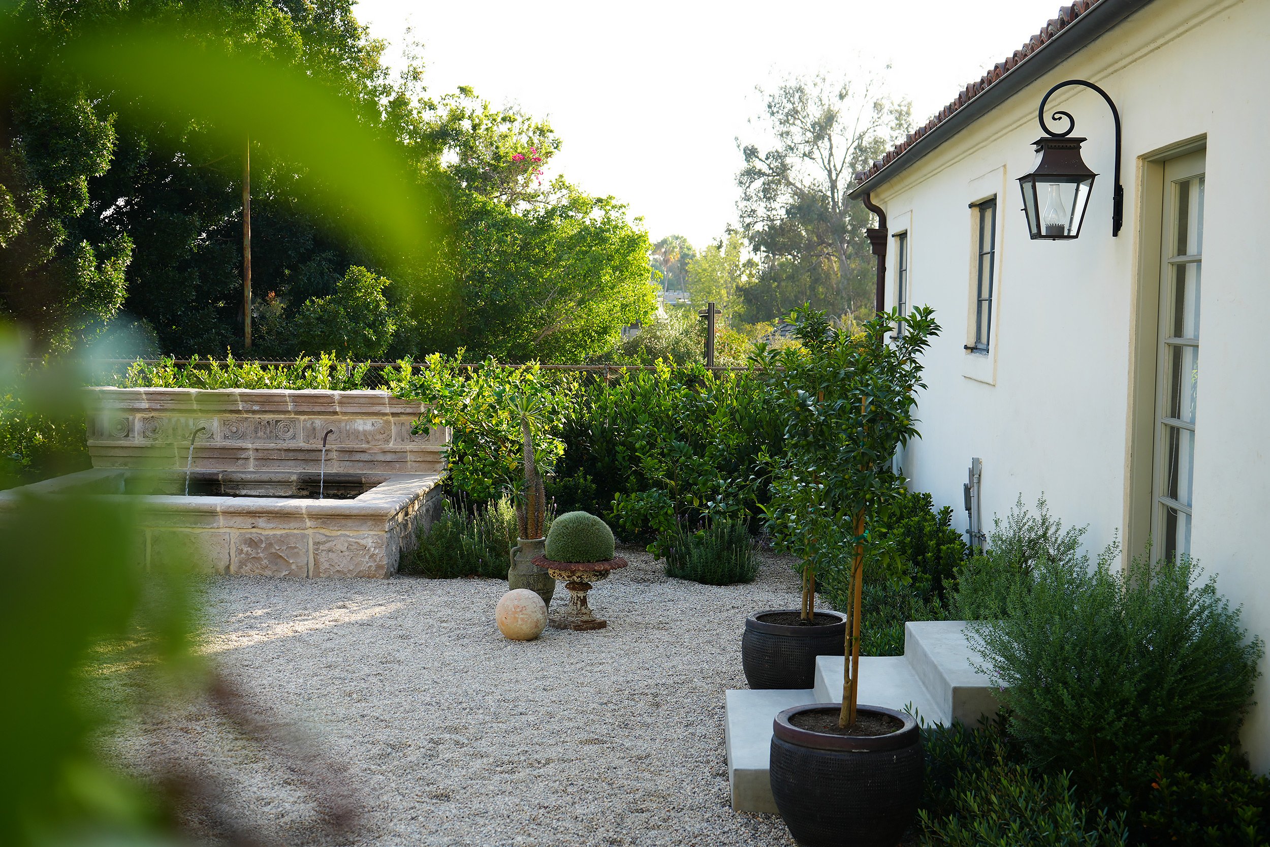 A serene outdoor patio area with a white house, potted plants, a stone fountain, and lush green trees in the background.