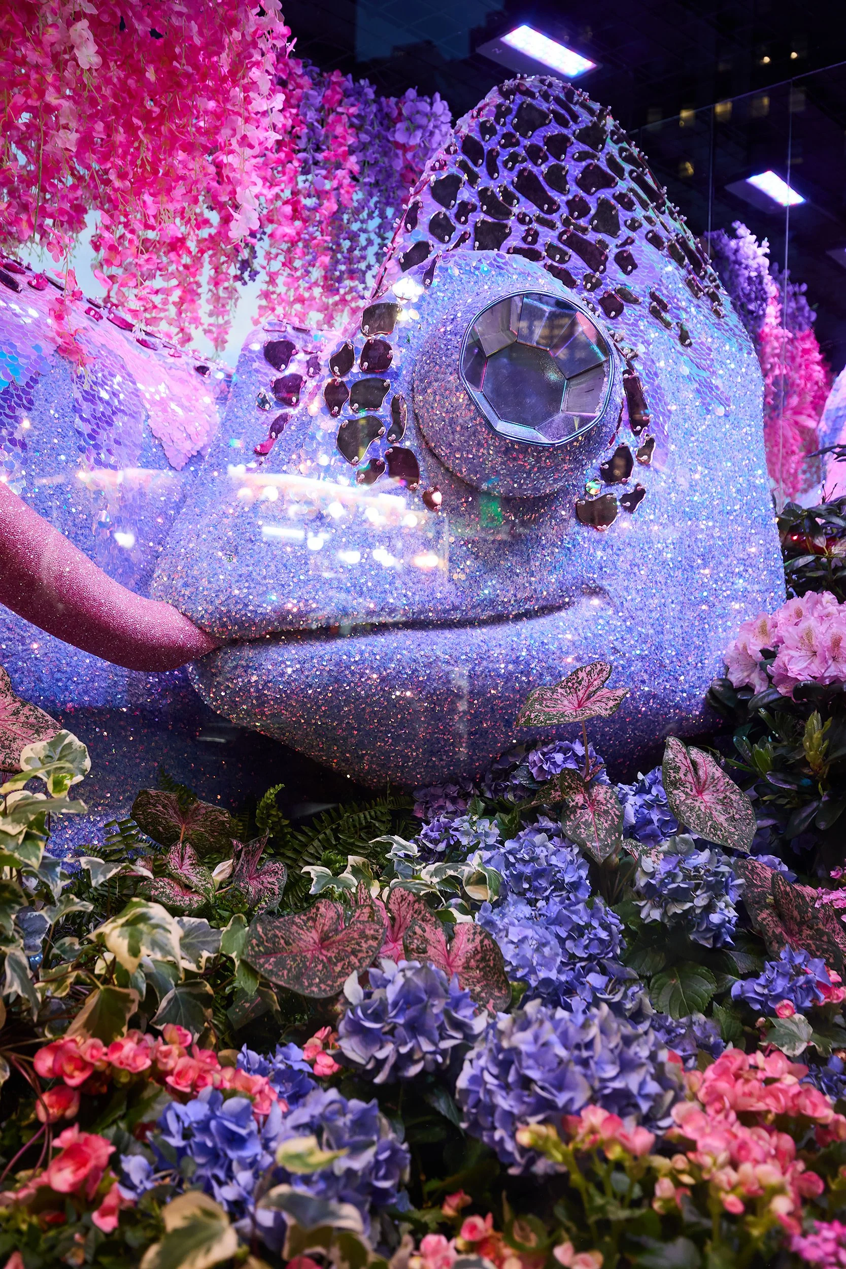 A large sculpture of a hand holding a camera, covered in glitter, surrounded by pink and purple flowers and foliage.