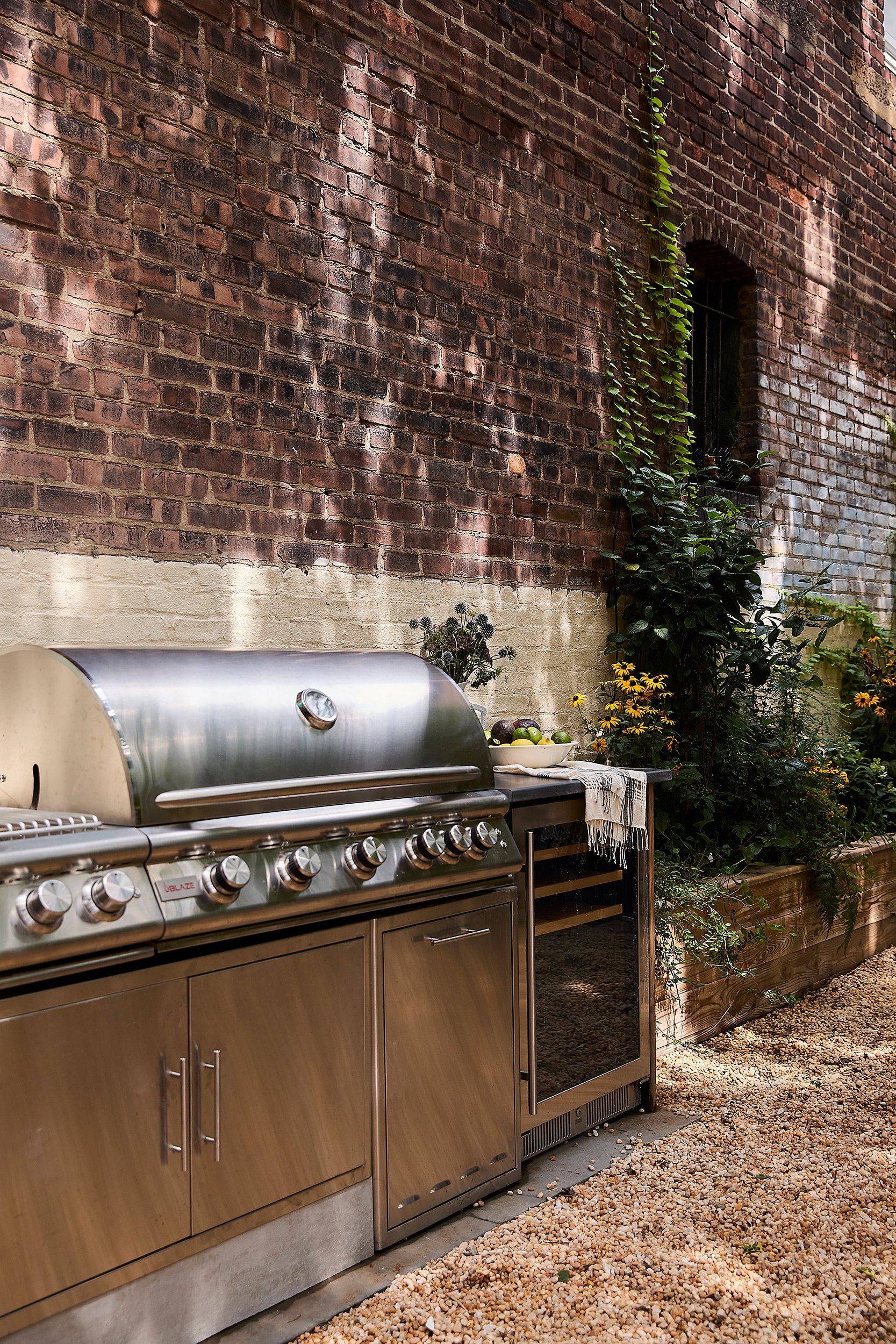 An outdoor kitchen setup with a stainless steel grill, a small mini fridge, a wooden countertop with a bowl of green apples, and various plants against a red brick wall.