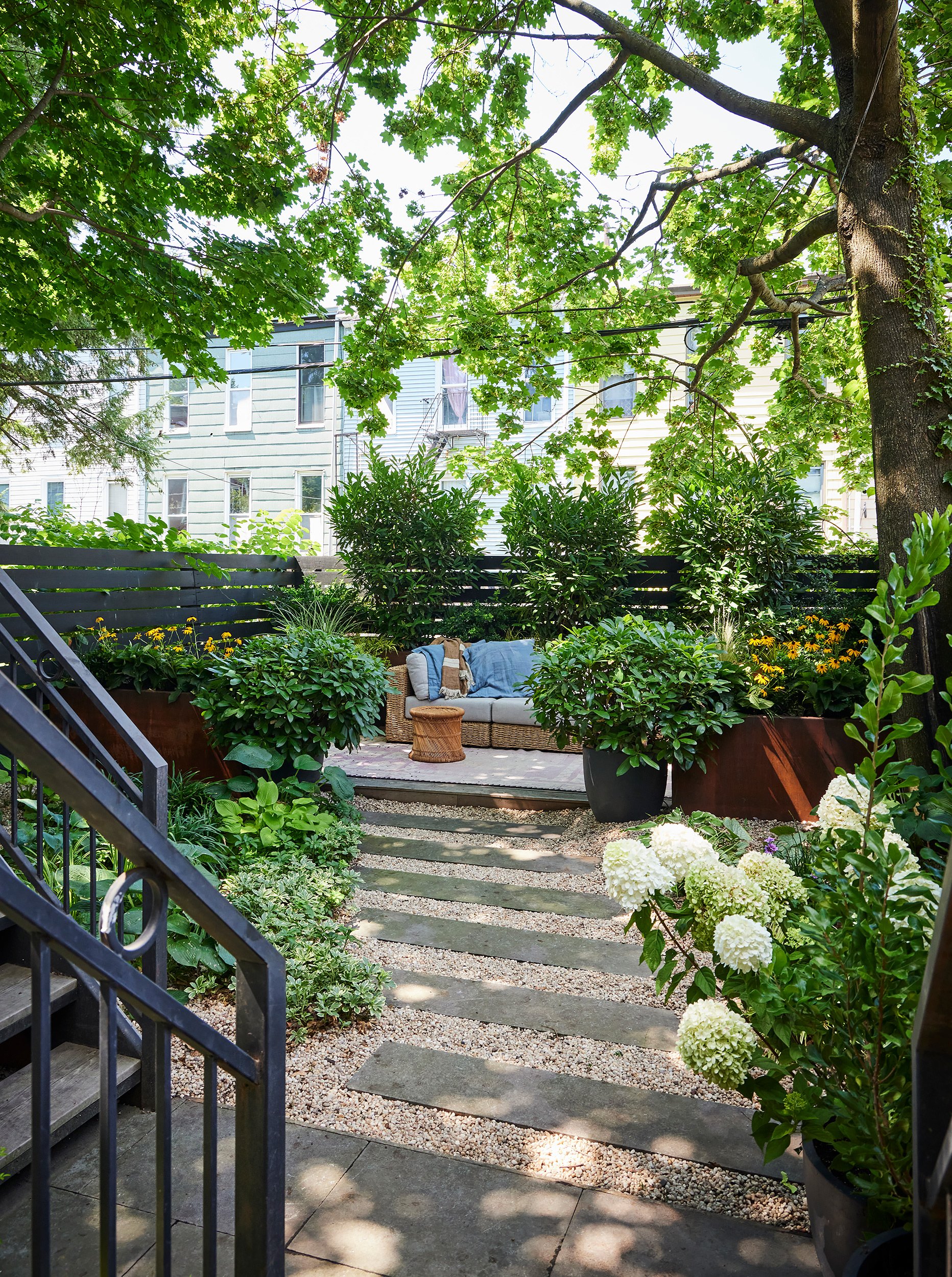 A cozy backyard patio with a seating area, surrounded by lush green plants and trees, with a pathway made of stone slabs on gravel leading to the seating area with an outdoor couch and small table.
