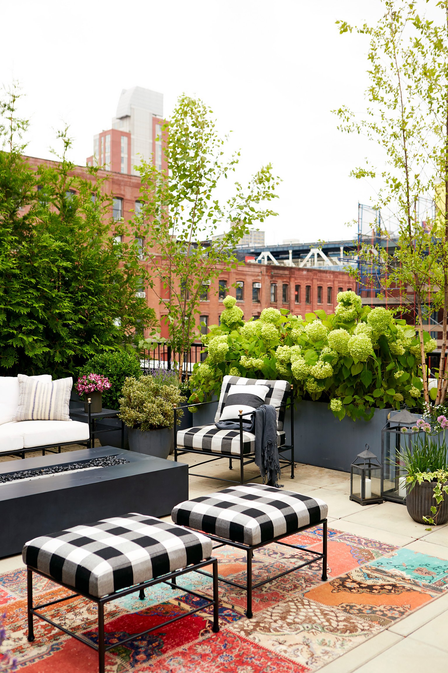City rooftop patio with outdoor furniture, plants, lanterns, and a patterned rug under an overcast sky.