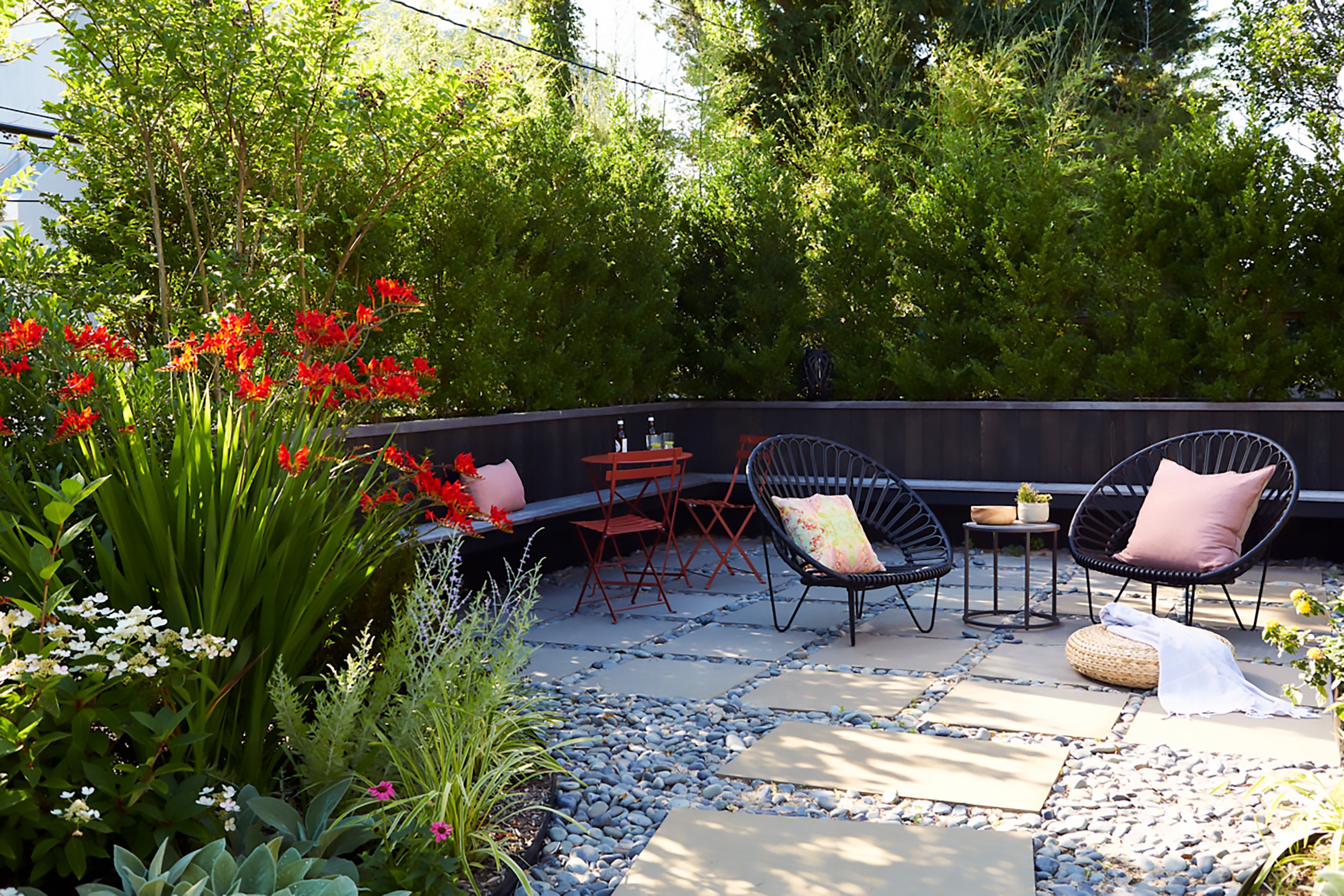 Outdoor patio with black circular chairs, pink pillows, a small black side table, a red table with two red chairs, surrounded by lush green plants and colorful flowers, with sunlight filtering through.