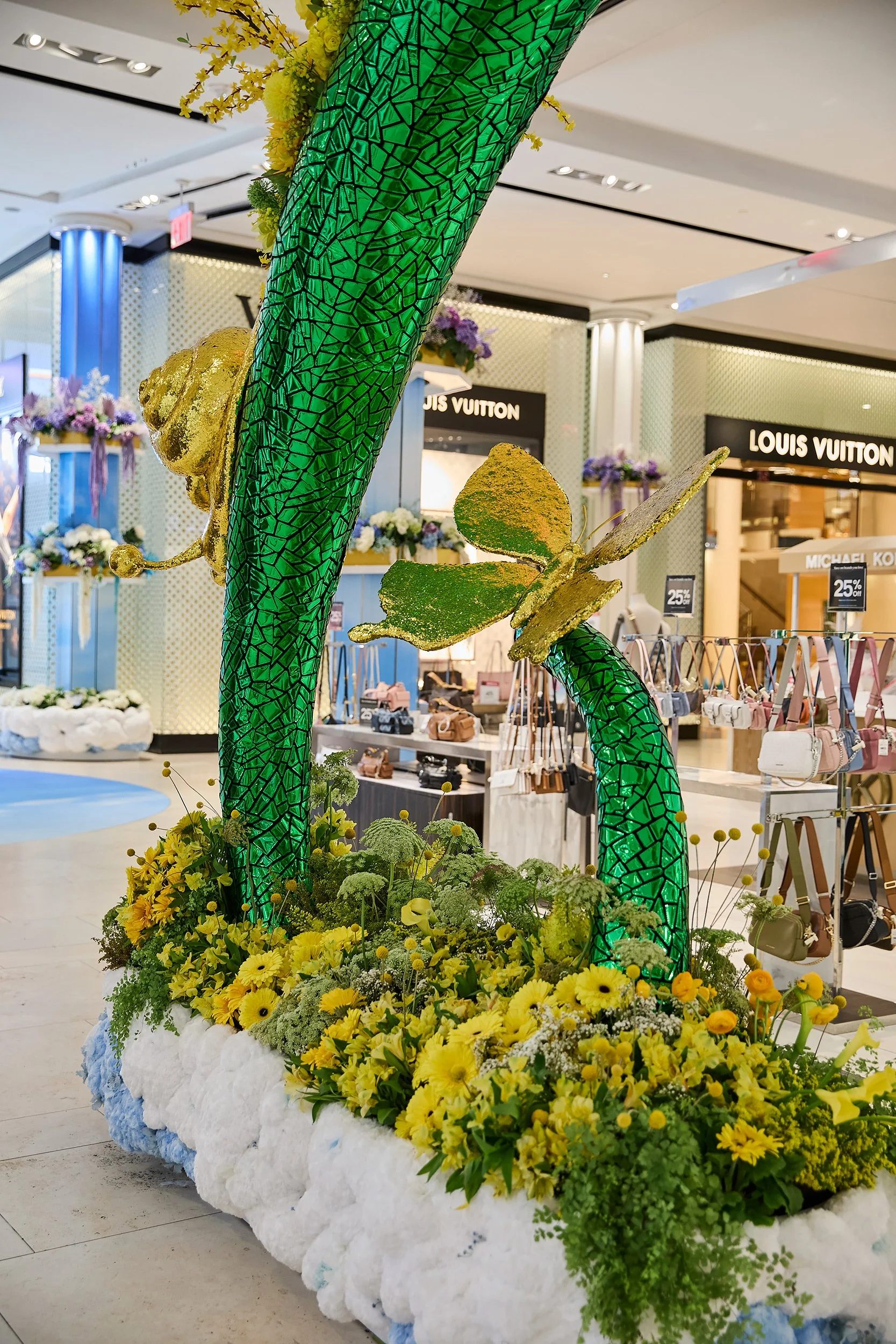 Decorative display with a large green mosaic arch, gold bee and butterfly figures, and yellow flowers, set inside a shopping mall with Louis Vuitton store in the background.