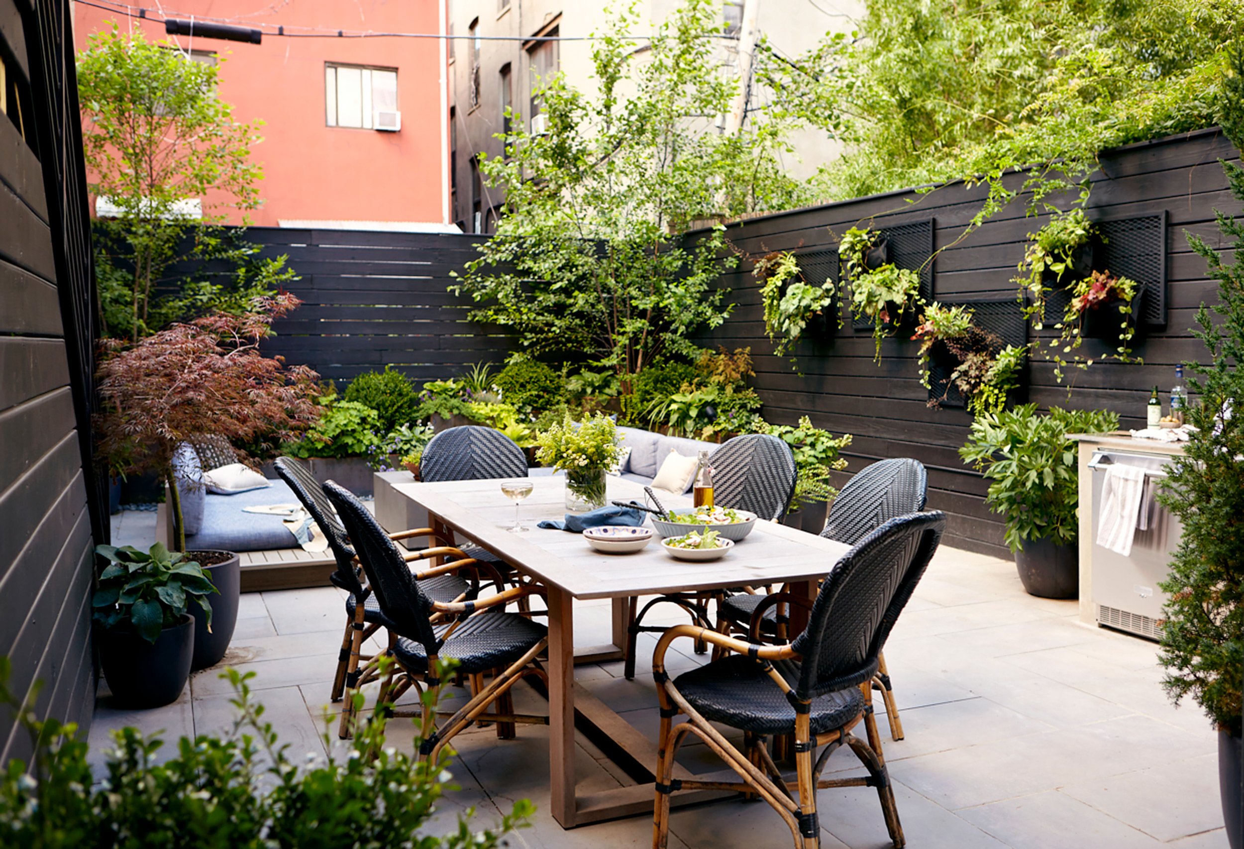 Outdoor patio with a dining table and six black chairs, surrounded by potted plants and greenery, with a black wooden fence and buildings in the background.