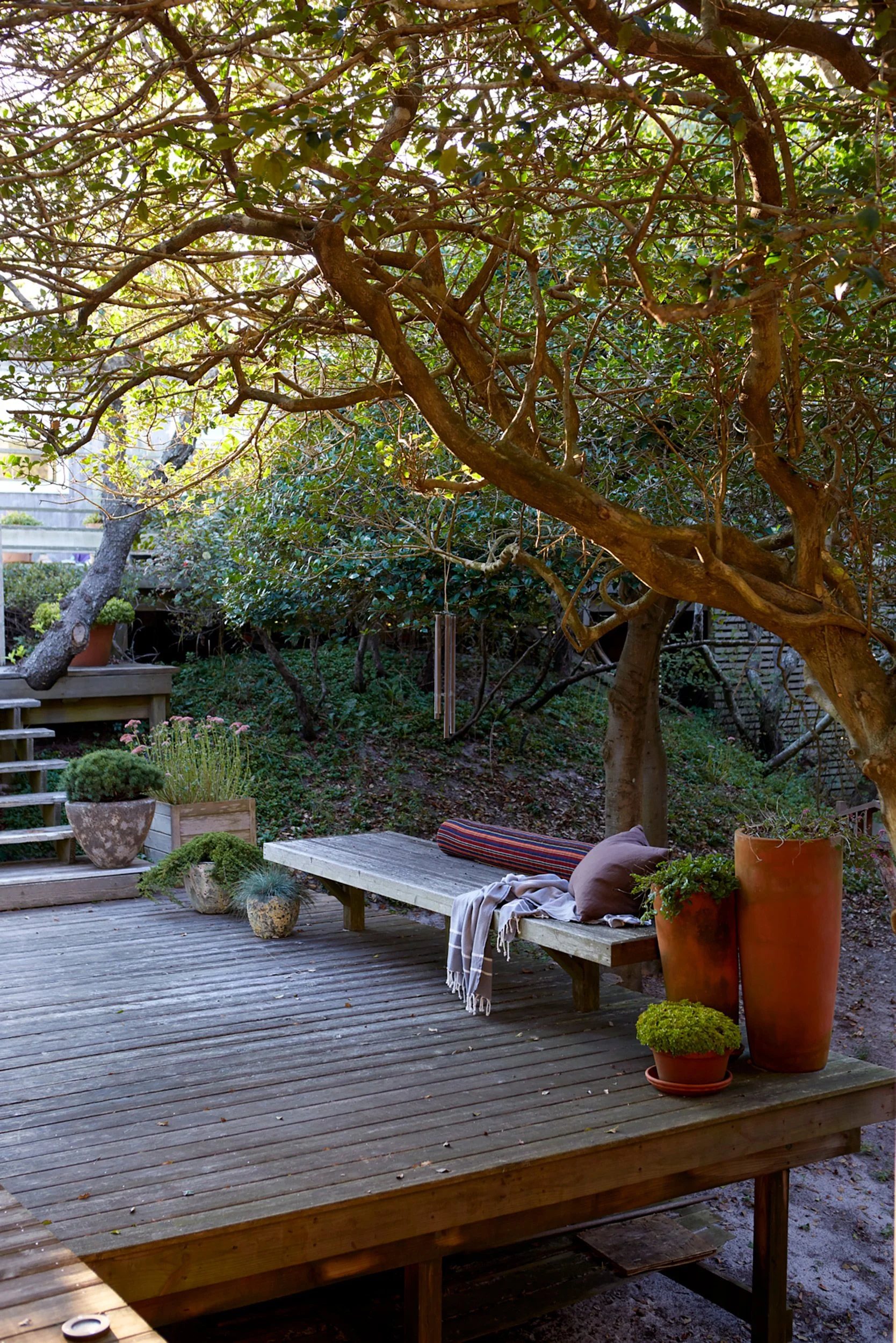 A backyard deck with potted plants, a bench with pillows and a blanket, surrounded by greenery and trees.