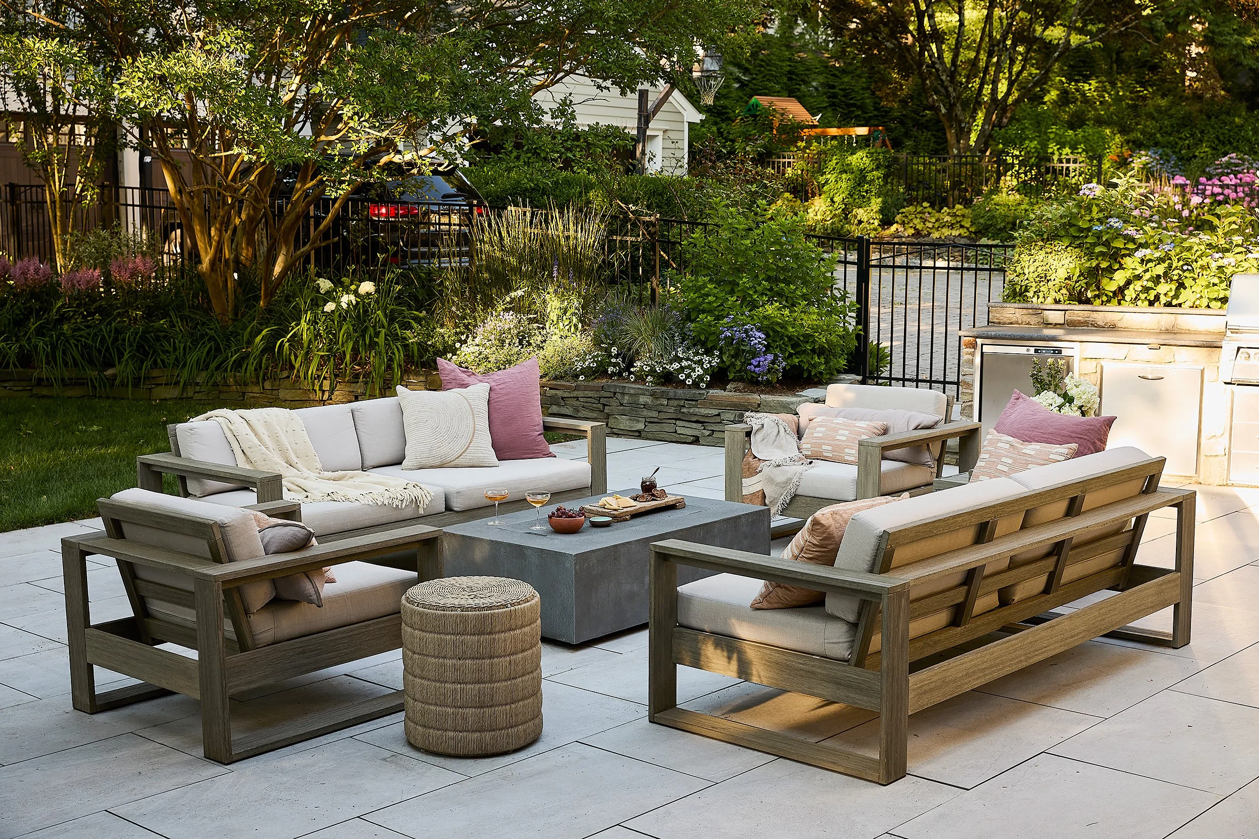 Outdoor patio with wooden furniture, cushioned seats, throw pillows, and a concrete coffee table, surrounded by garden plants and trees.
