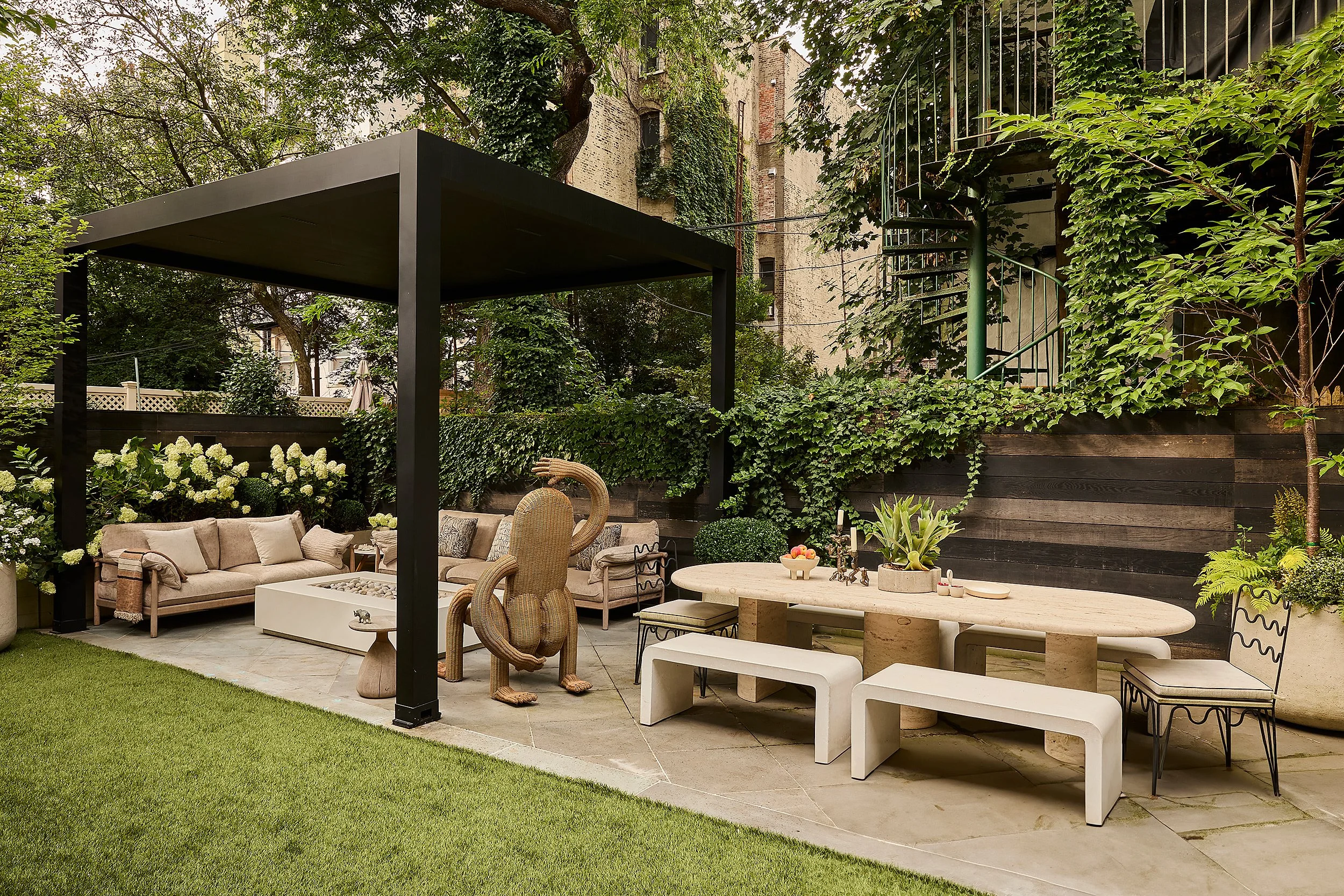 Outdoor patio with beige sofa, white coffee table, stone dining table, benches, and chairs under a black canopy, surrounded by green plants and trees in an urban garden.