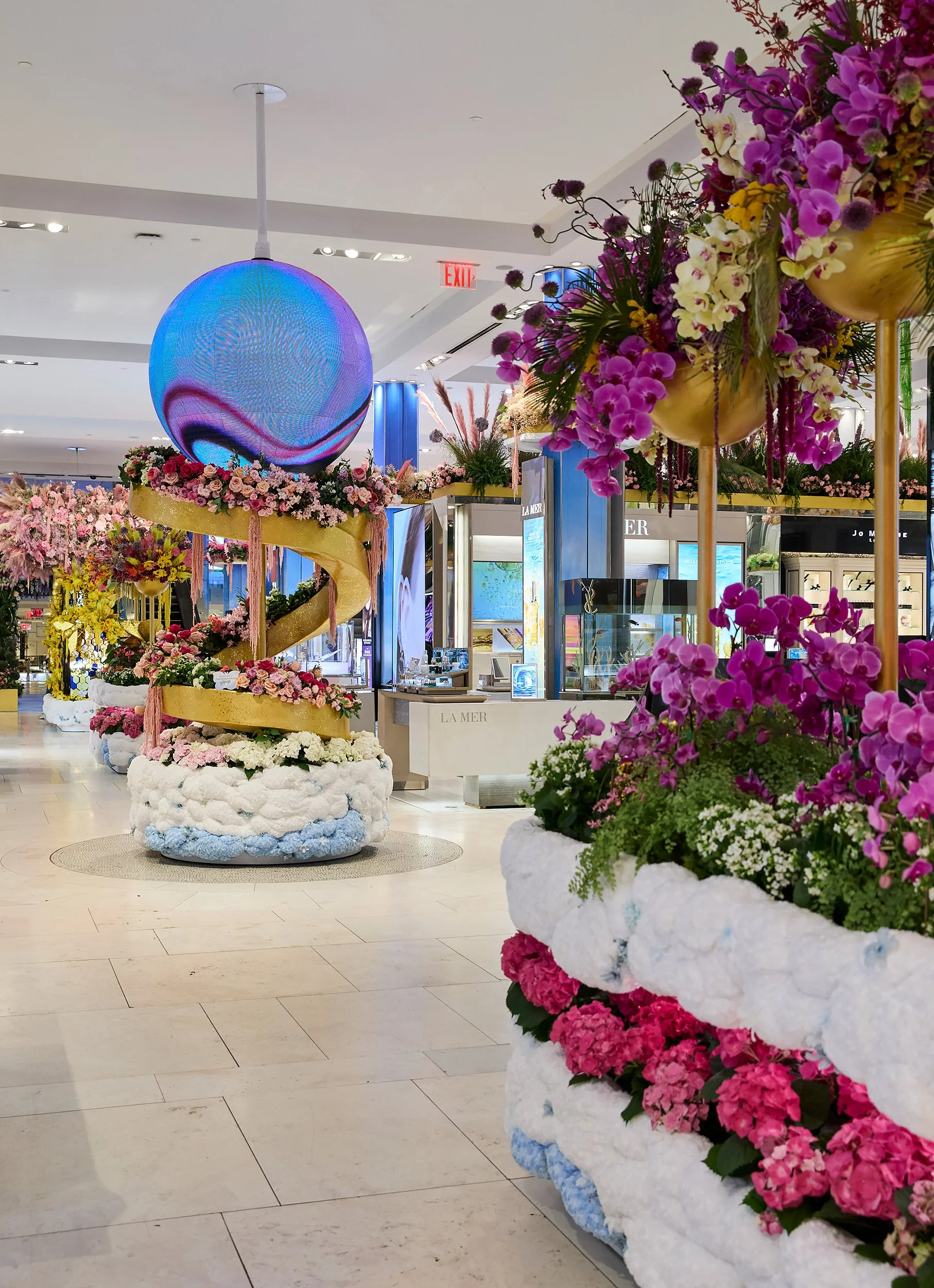 Decorative floral display inside a shopping mall, featuring pink, purple, and white flowers arranged in large planters, with a blue circular LED light and a yellow spiral structure as central decorations.