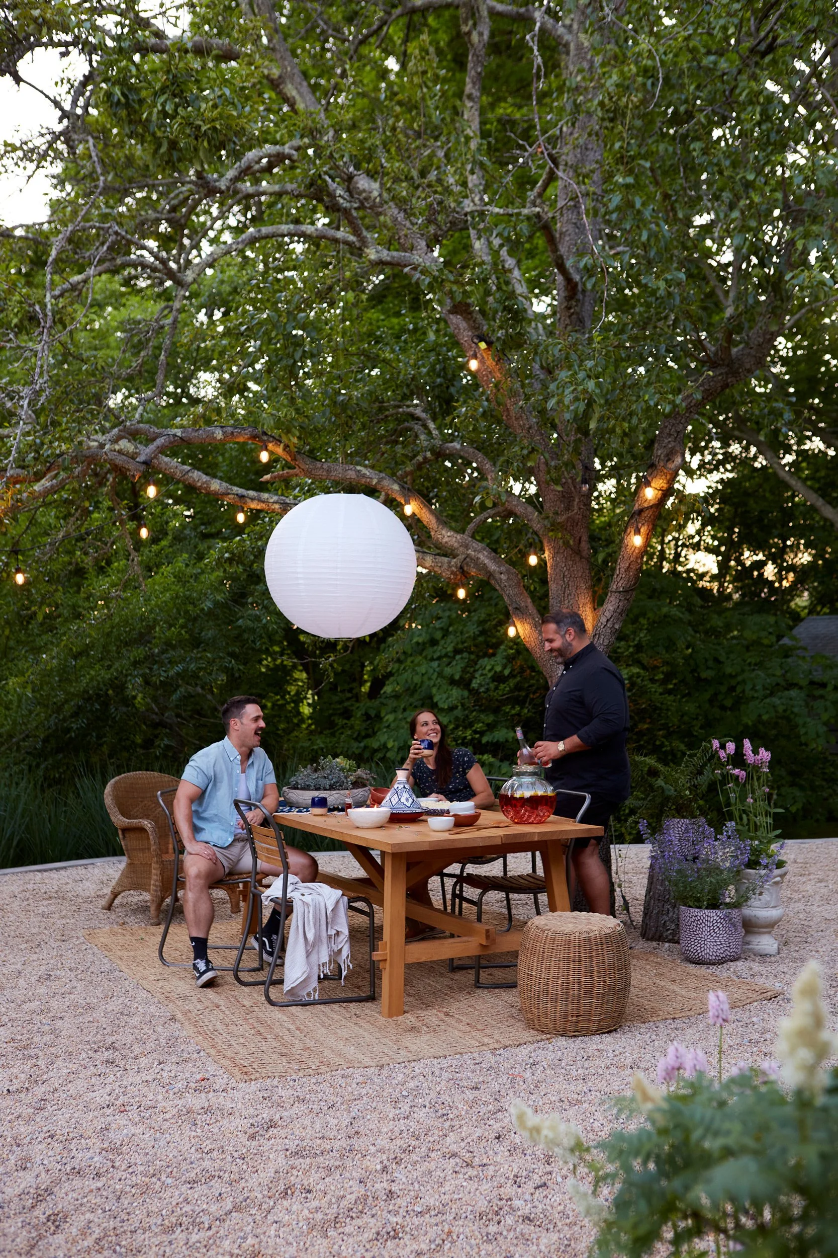 People having outdoor dinner under a tree decorated with string lights and a large paper lantern, surrounded by potted plants.