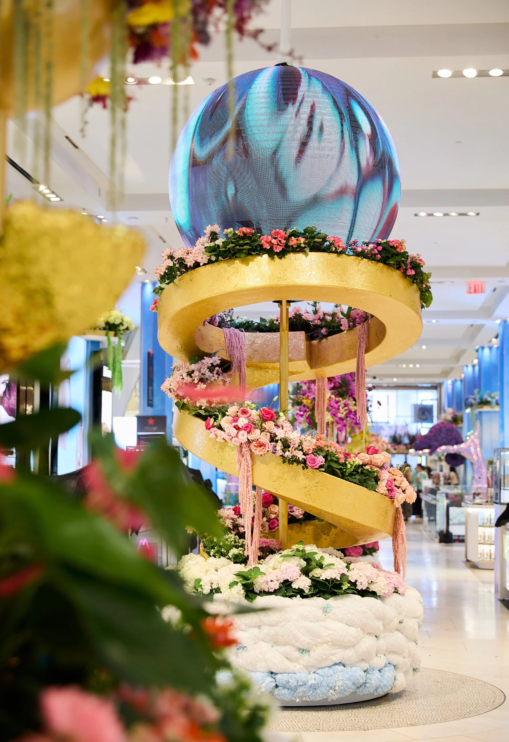 A decorative art piece featuring a spiral yellow platform with pink and white flowers, topped with a large, reflective solar ball in a mall or exhibition space.