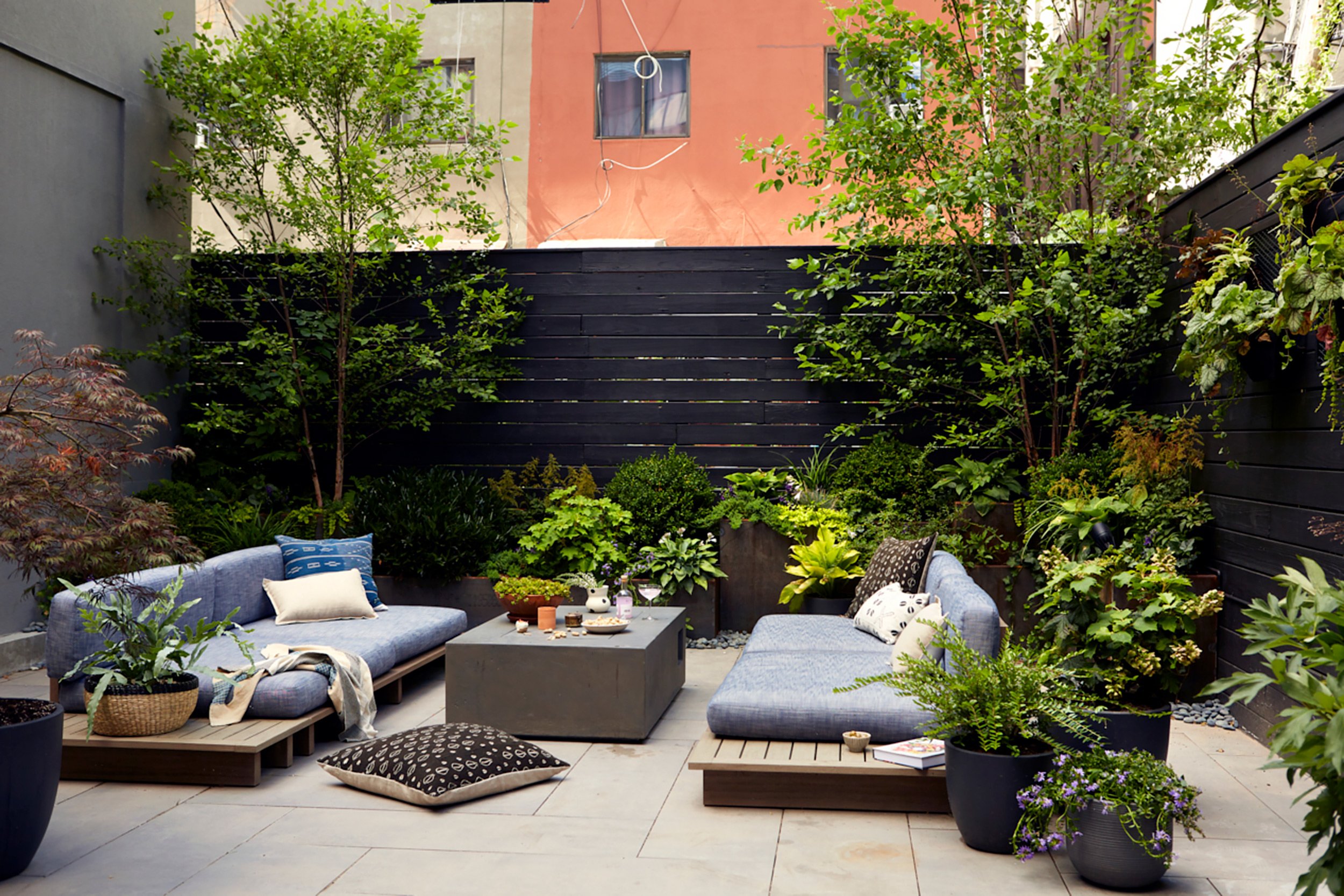 A cozy outdoor patio with two seating areas, surrounded by lush green plants and trees, enclosed by black wooden fencing, with a concrete coffee table in the center and colorful cushions on the couches.