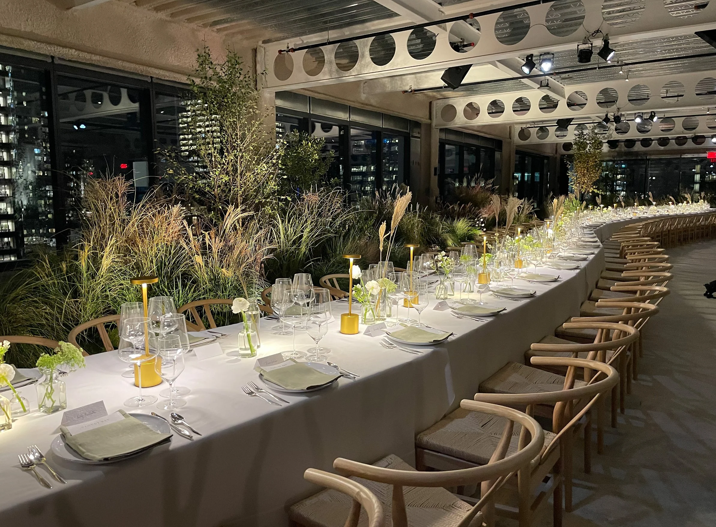 A long banquet table set for a formal dinner, decorated with small vases of white flowers, several wine glasses, and yellow lamps, with modern chairs and a city skyline visible through large windows in the background.