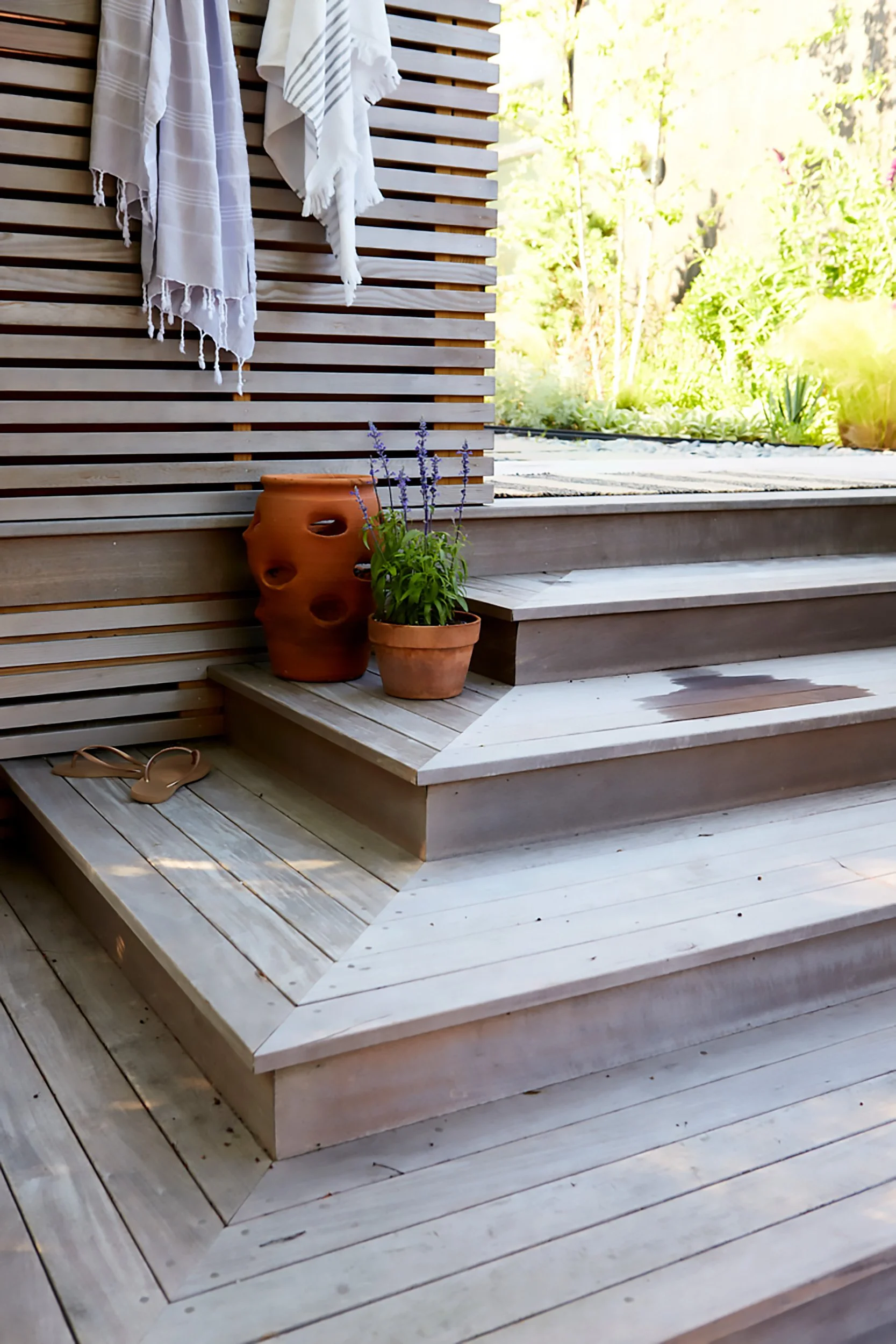 Wooden outdoor stairs with potted lavender and a large ceramic vase on the side, hanging towels on a wooden slat wall, with lush greenery in the background.