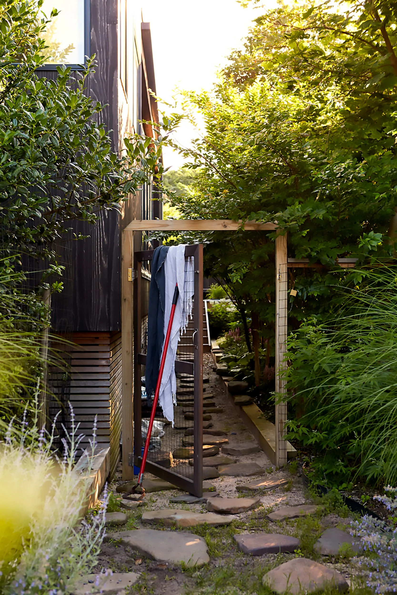 A garden pathway with stepping stones leading to a wooden gate, surrounded by lush green plants and trees, with sunlight coming through the foliage.