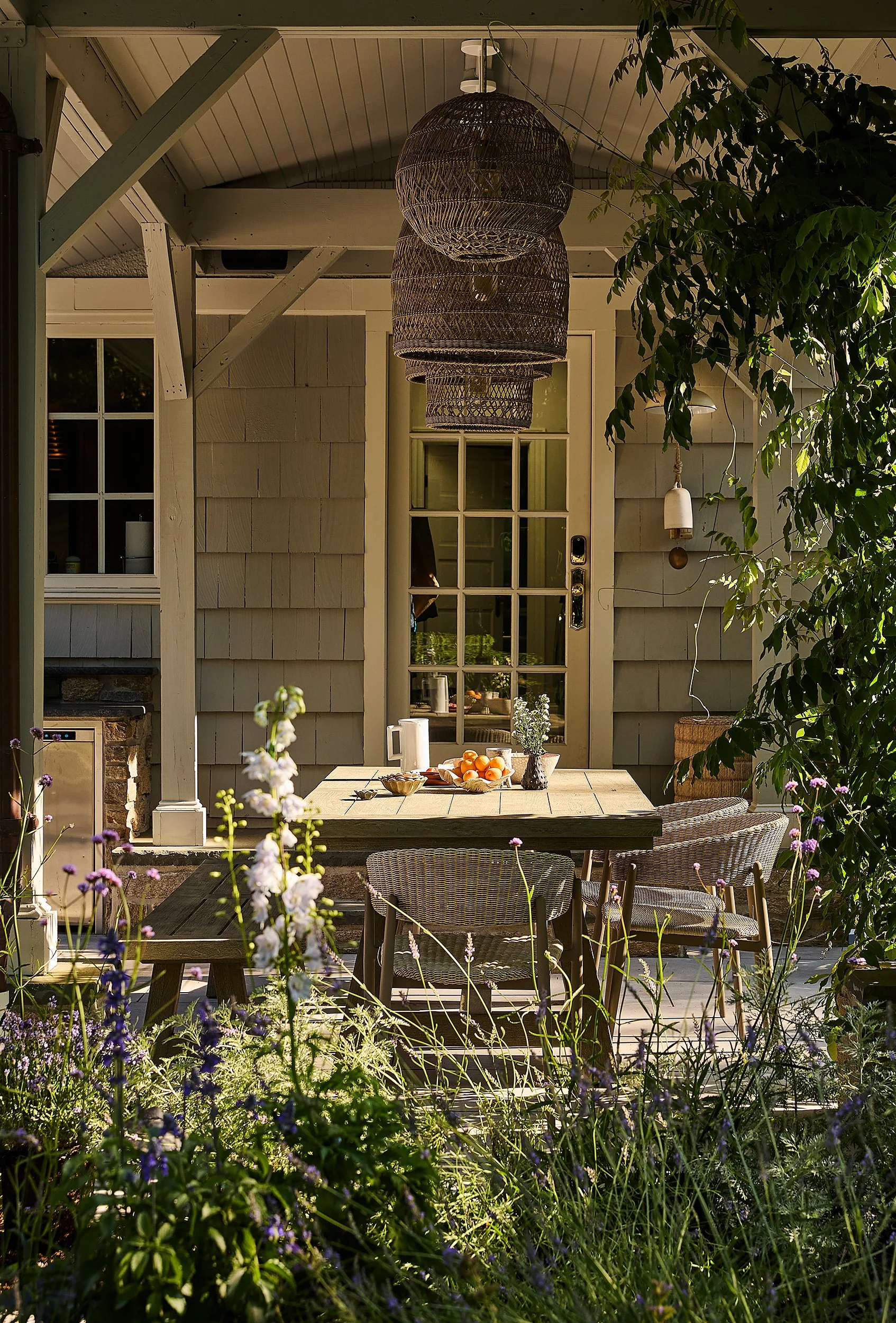 Outdoor porch with a wooden table, wicker chairs, hanging pendant lights, and surrounding lush green plants and flowers.
