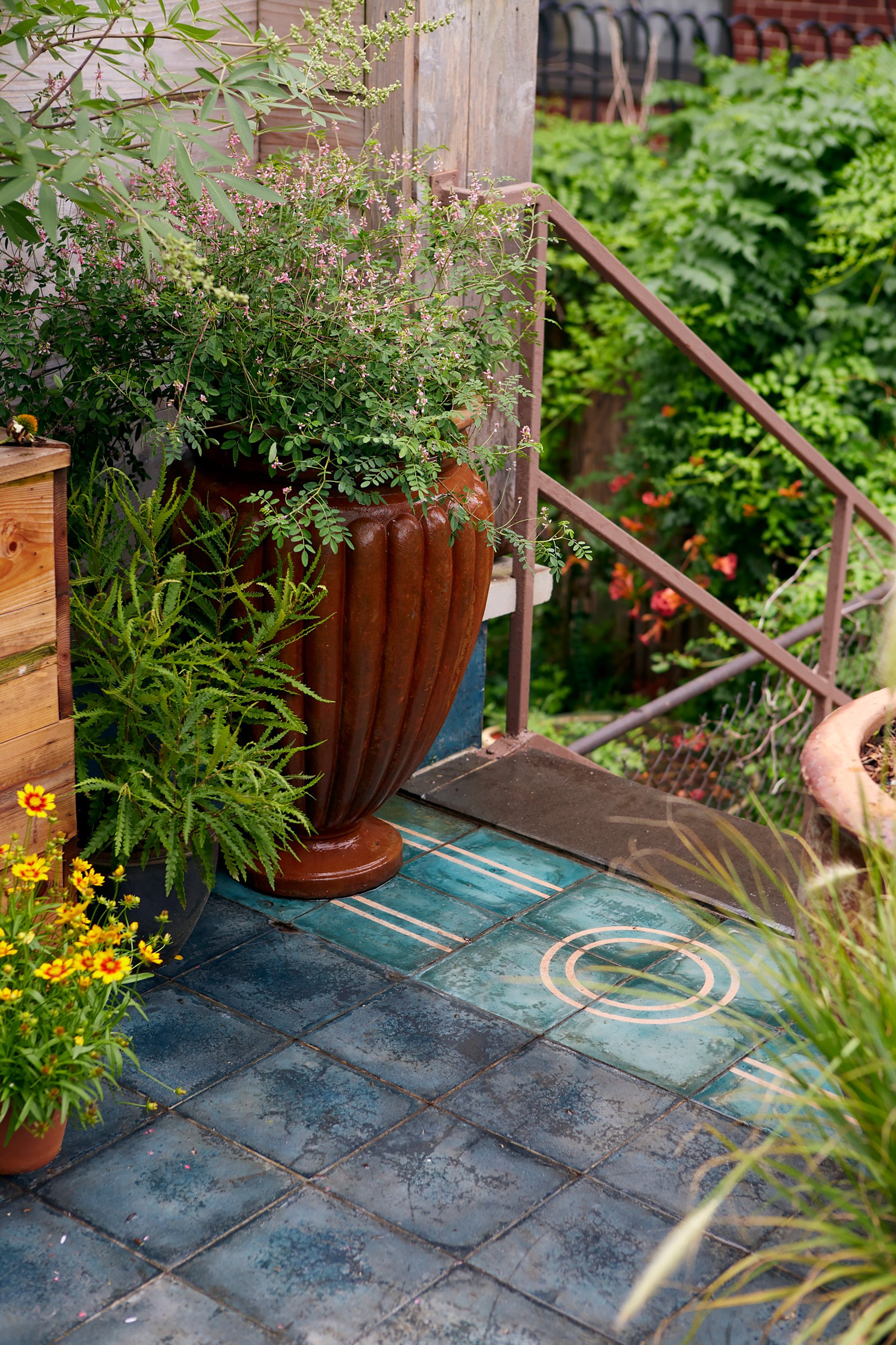 A cozy outdoor porch with potted plants, including a large terracotta vase with a green and pink flowering plant, and a smaller black pot with ferns, set on a tiled surface with a circular decorative pattern. Green foliage is visible in the background.