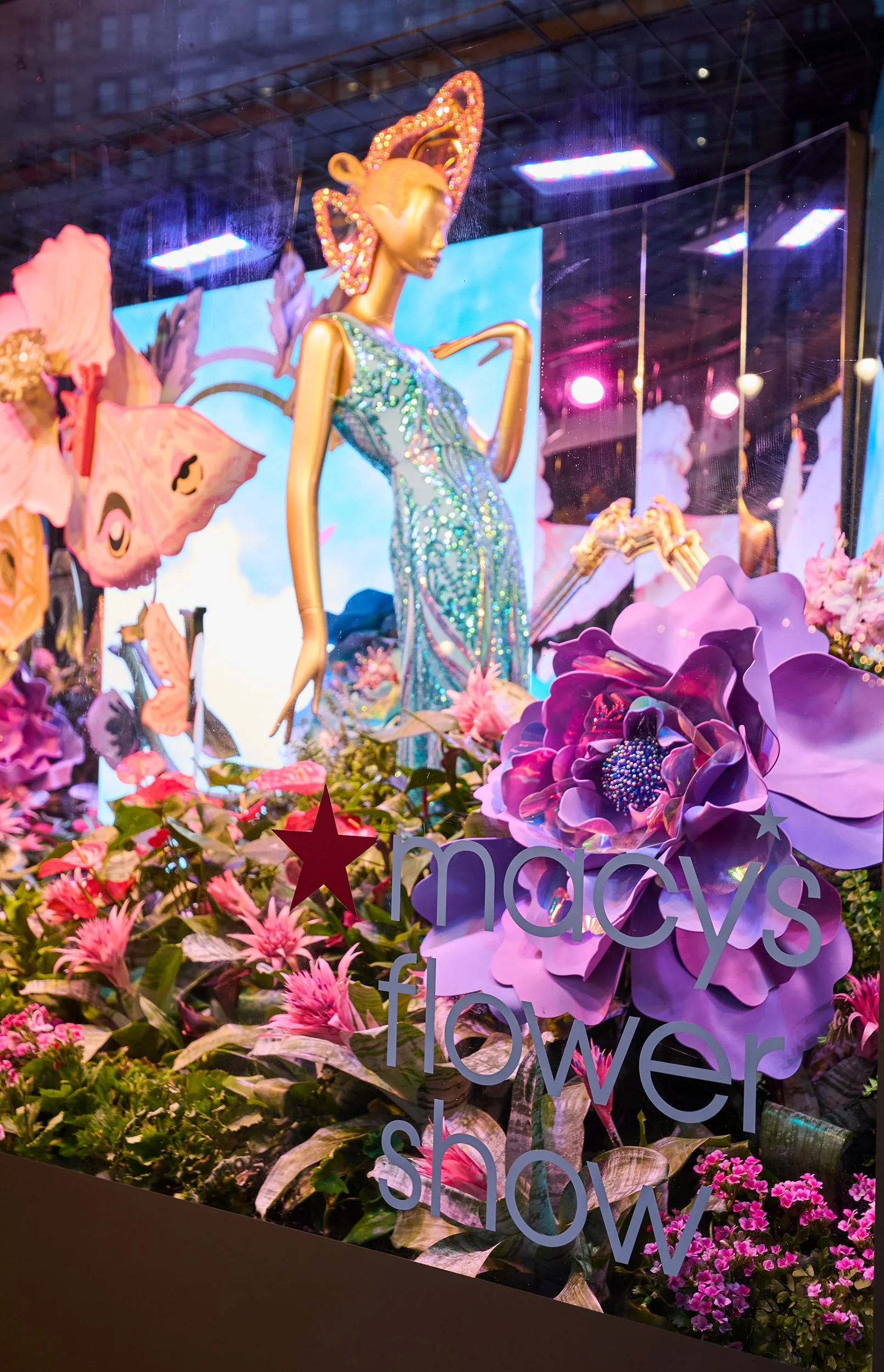 Store window display featuring a glamorous mannequin in a sparkling dress with a butterfly headpiece, surrounded by large pink and purple flowers and butterflies, with a sign reading 'Macy's Flower Show'