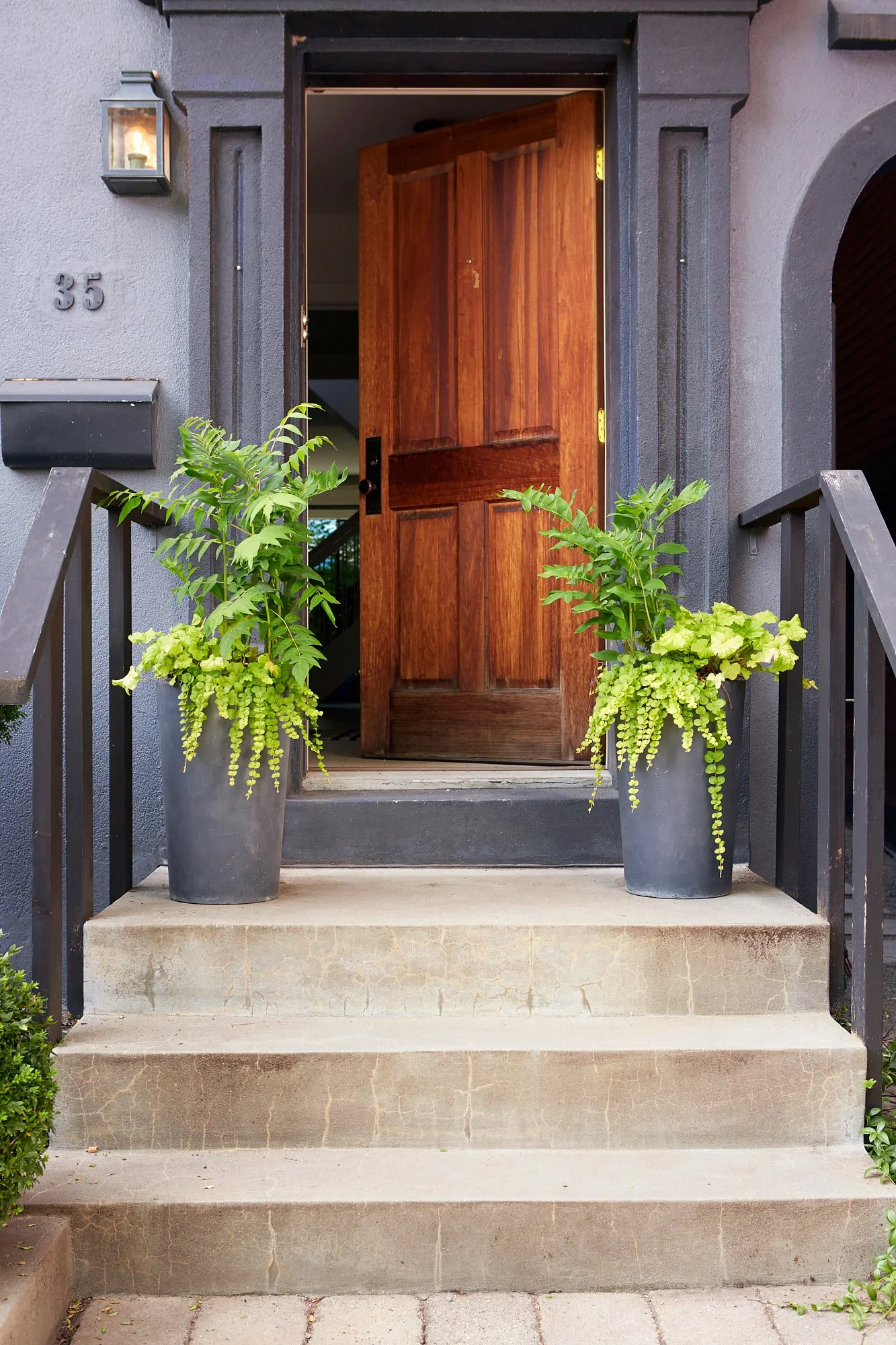 Front porch with open wooden door, potted plants on steps, gray exterior, house number 35, black railing, lantern-style light next to door.