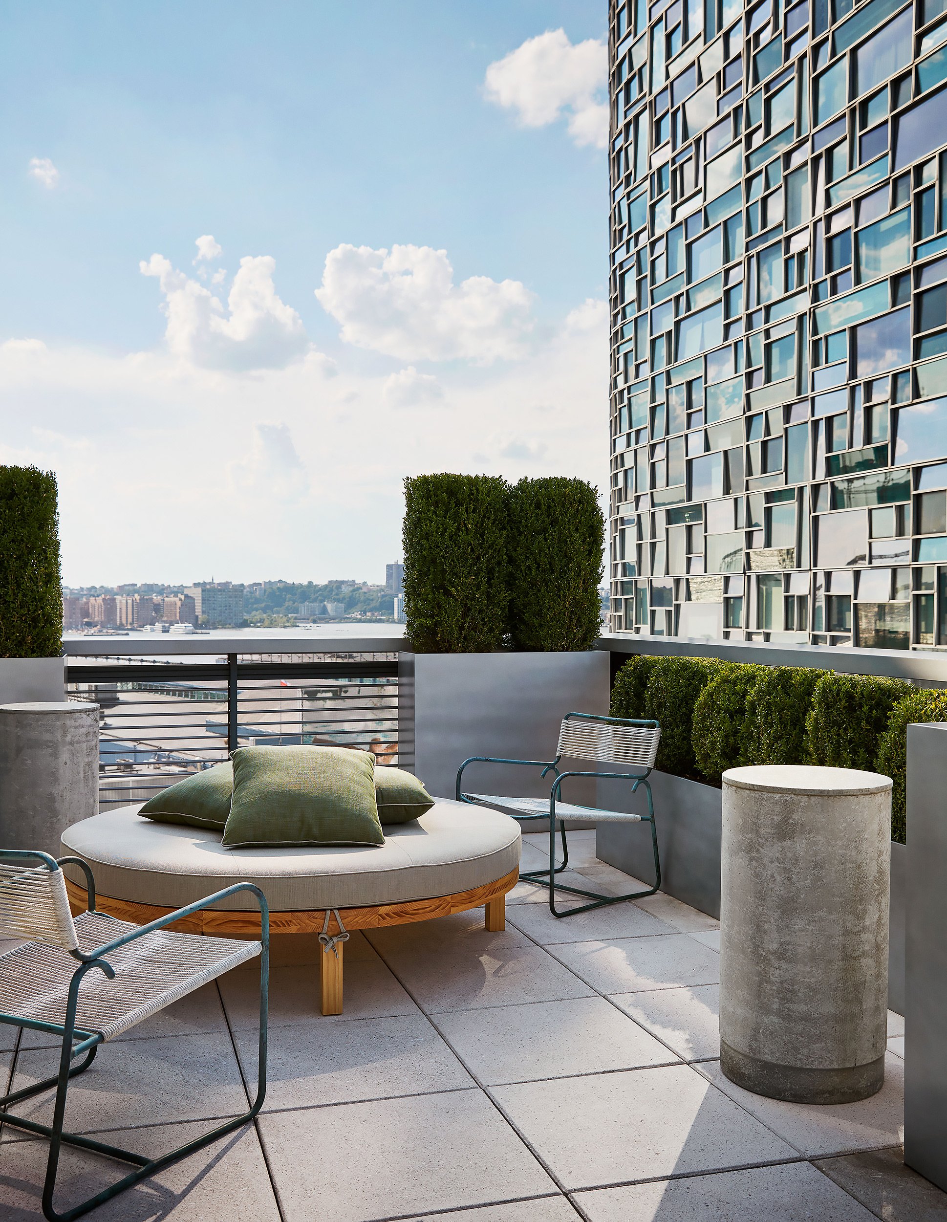 Rooftop patio with round outdoor daybed with green pillows, surrounded by patio chairs, large concrete planters, trimmed hedges, glass building, and city skyline in the background under partly cloudy sky.