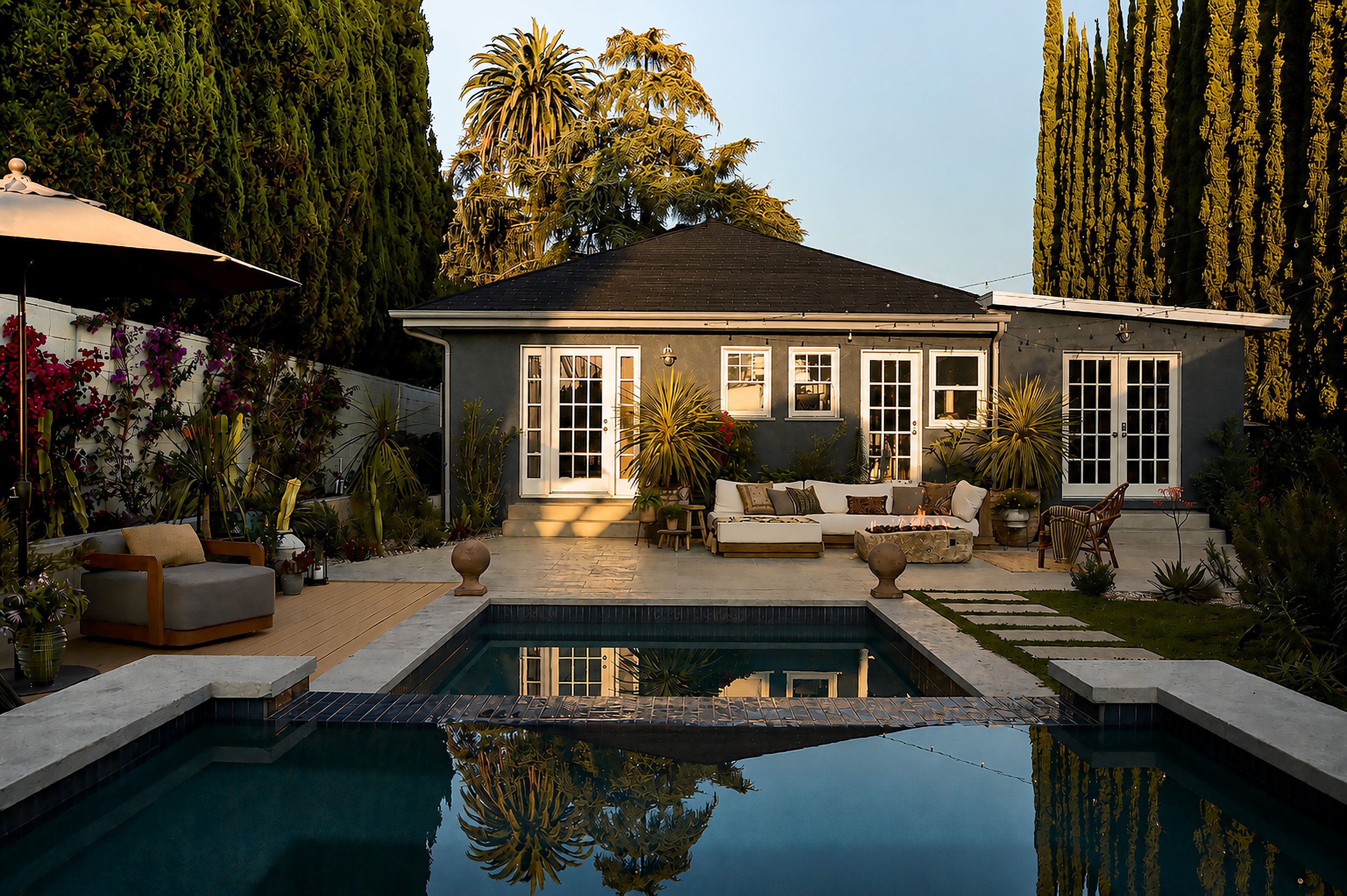 A backyard patio with outdoor furniture, potted plants, and a swimming pool reflecting a small gray house and trees in the background, during sunset.