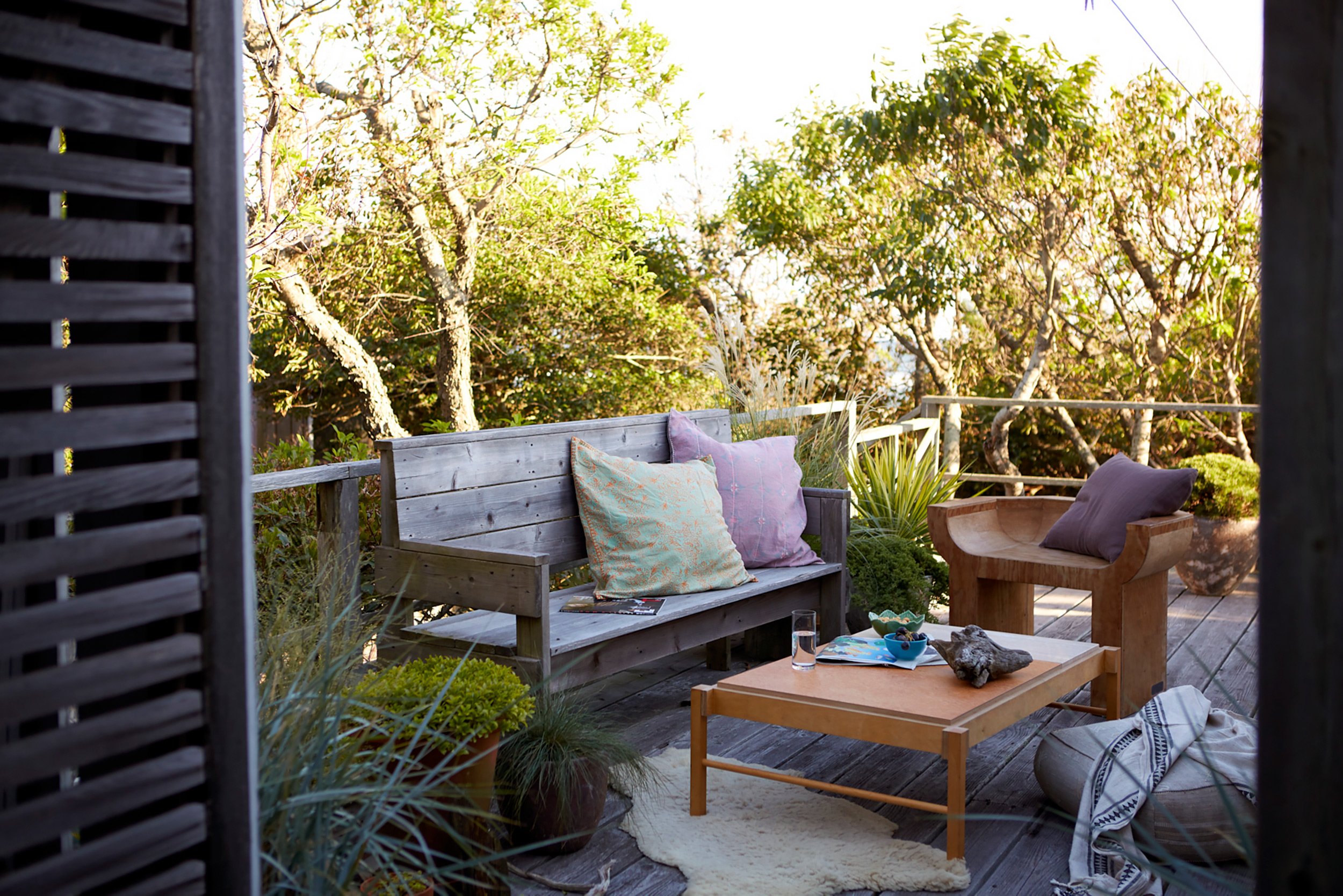 Cozy outdoor patio with wooden furniture, colorful cushions, potted plants, and a scenic view of trees and sky in the background.